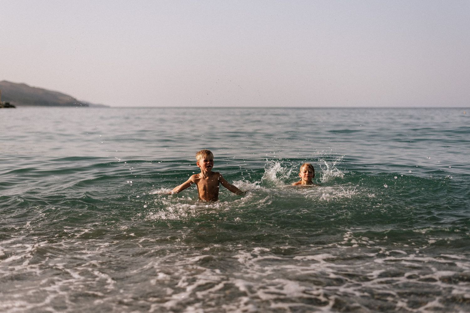 Two children splashing and swimming in calm turquoise ocean water near a rocky coastline on a sunny day.