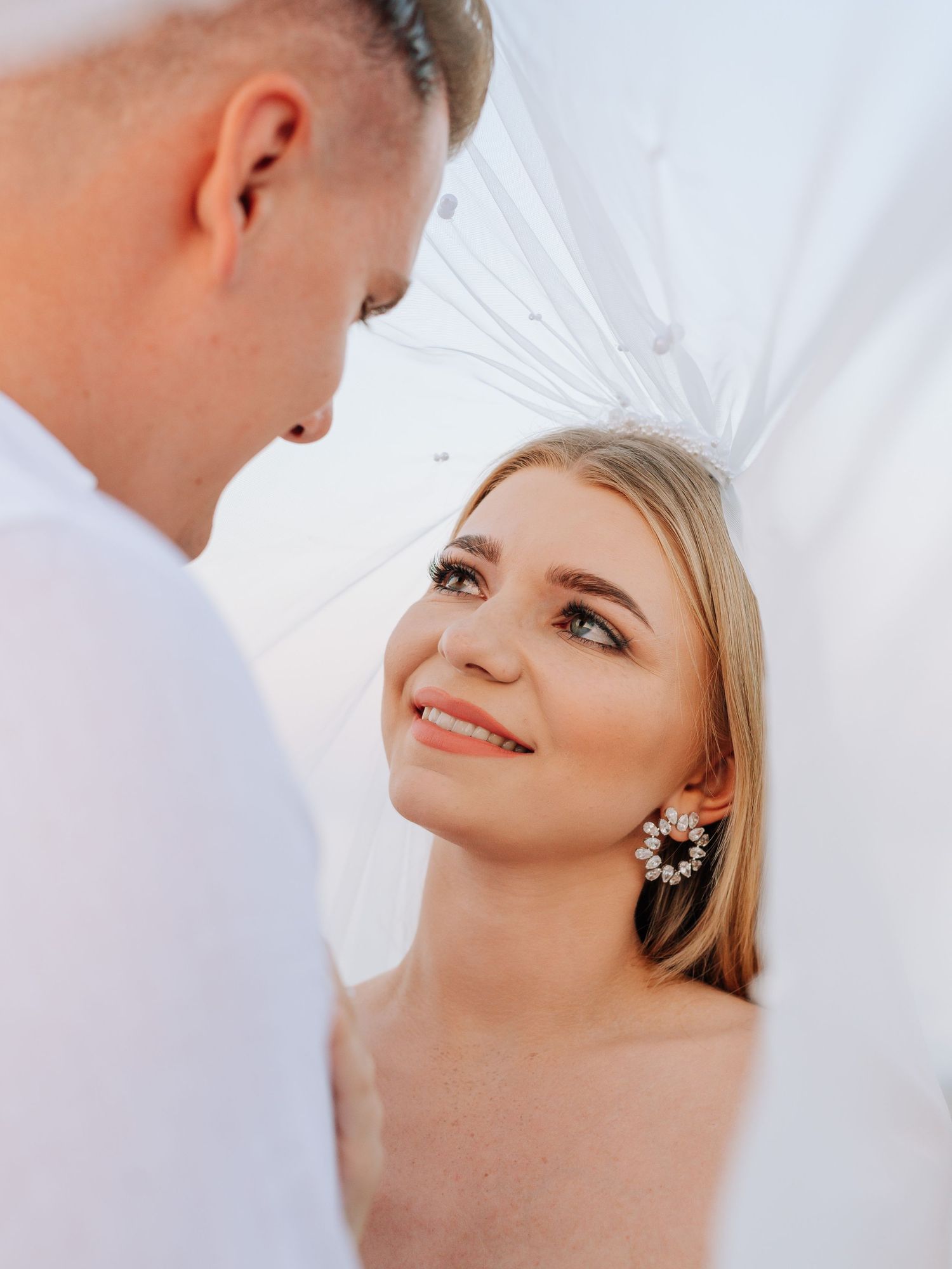 Smiling blonde bride wearing a veil and crystal earrings gazing at her groom.