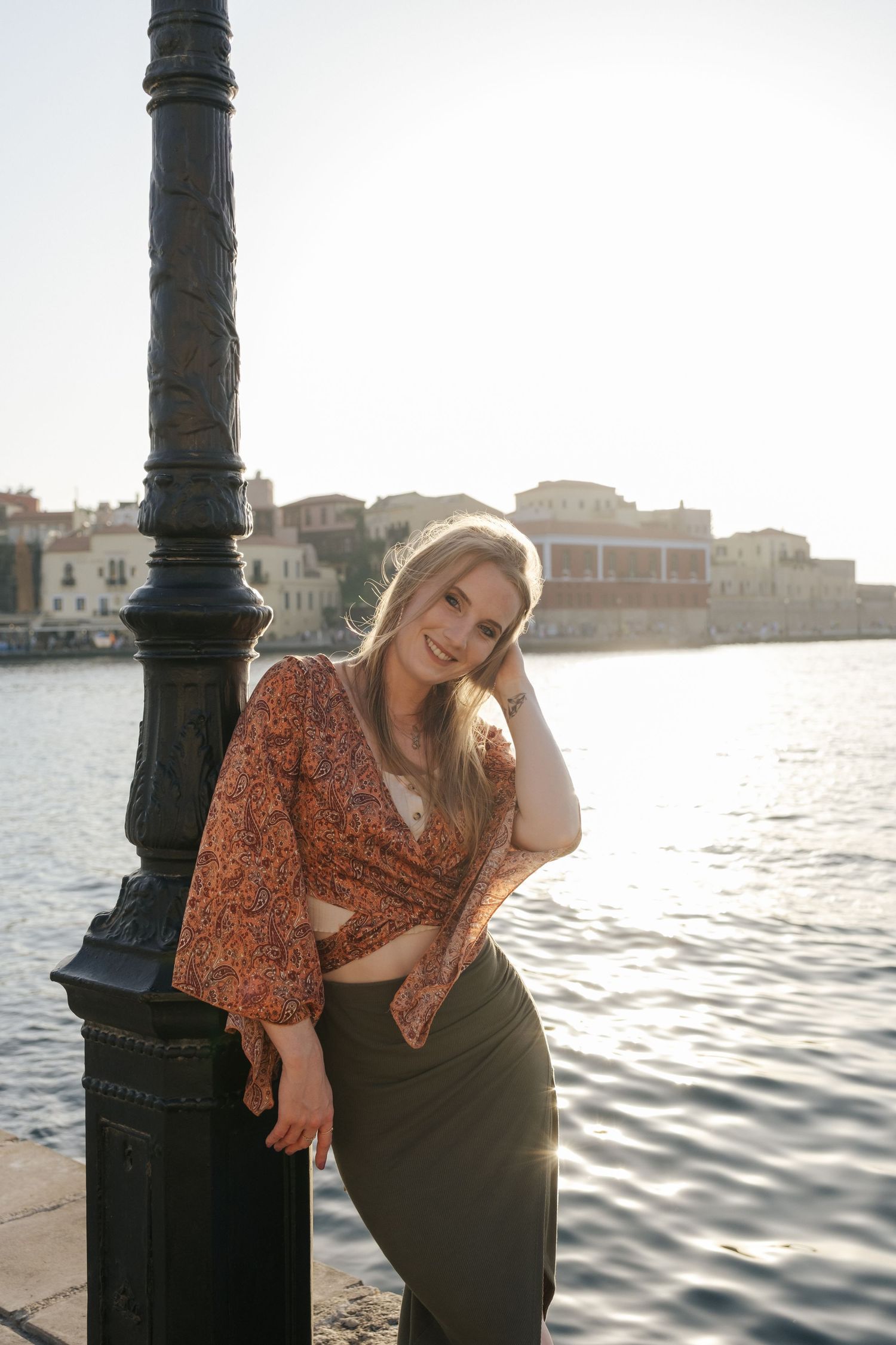 Smiling blonde woman in floral crop top and olive skirt leaning on waterfront lamppost.