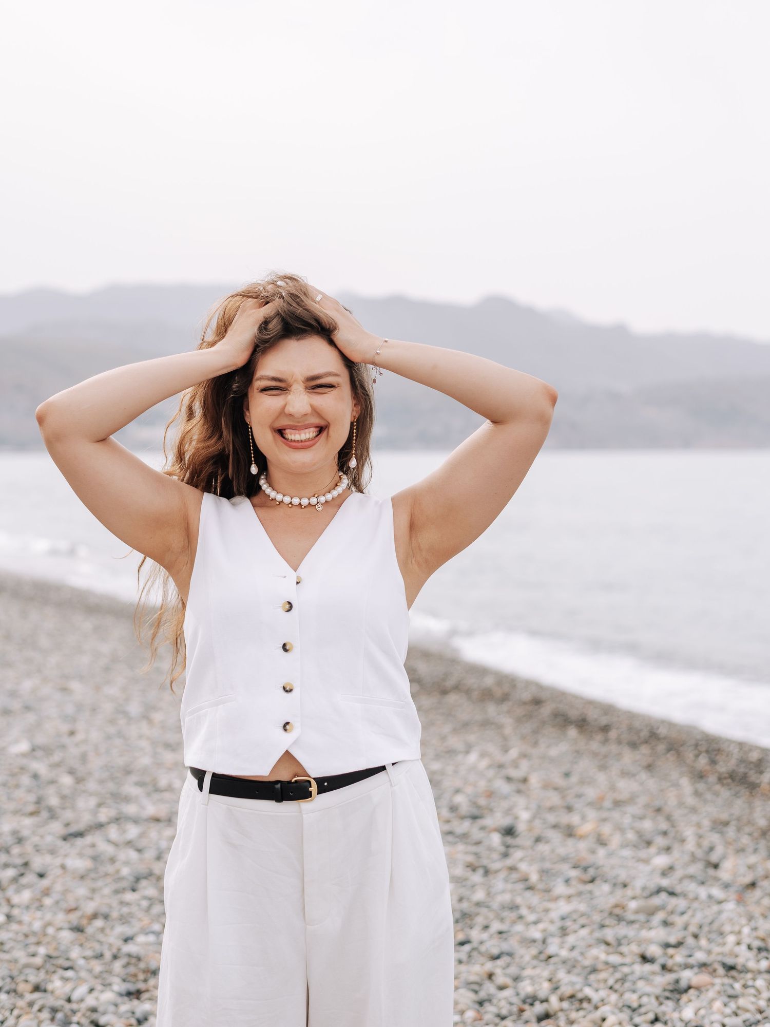 Smiling woman in white vest top and wide-leg pants with pearl necklace posing on pebble beach.