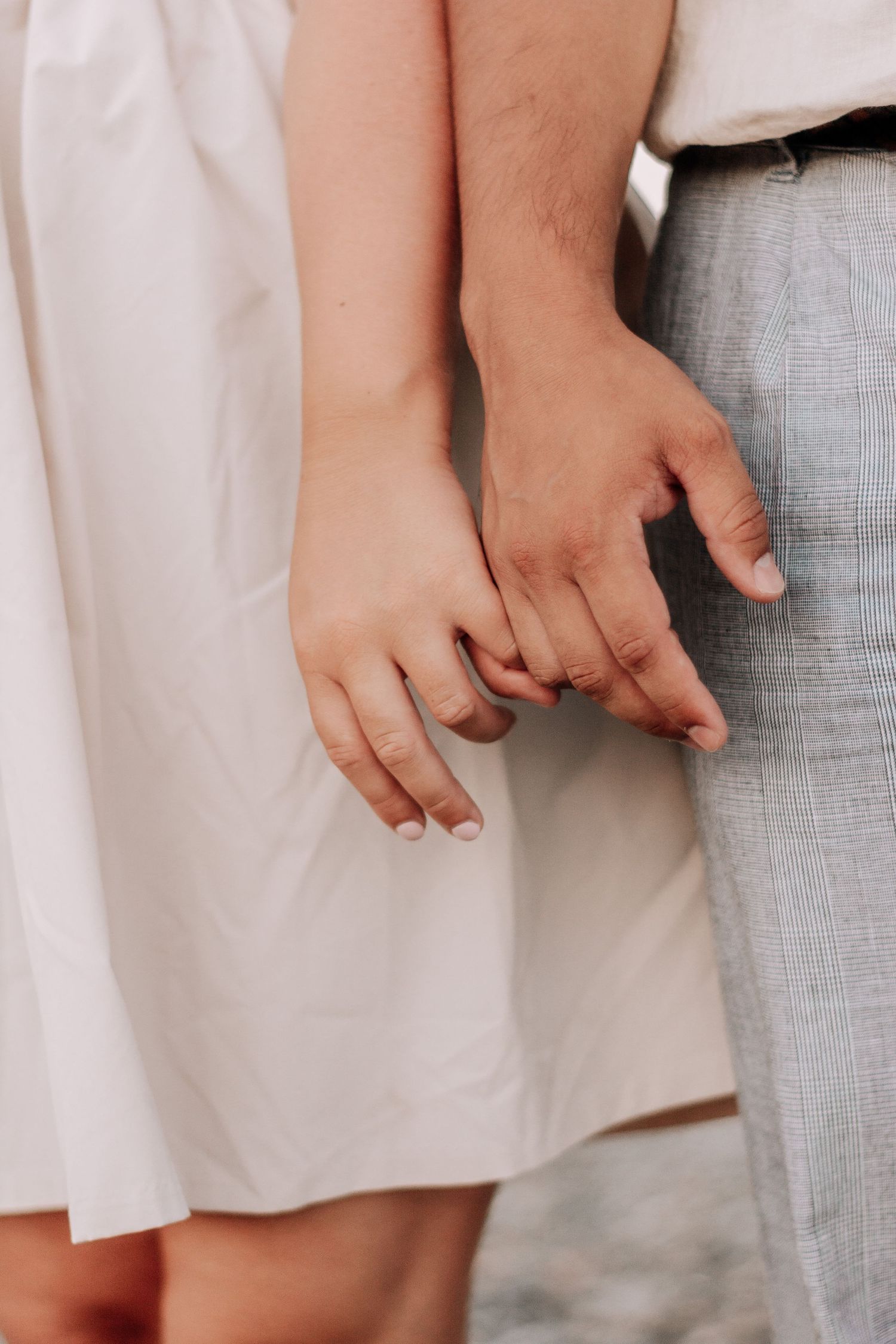 Close-up of a couple gently holding hands, woman in white dress and man in gray plaid trousers.