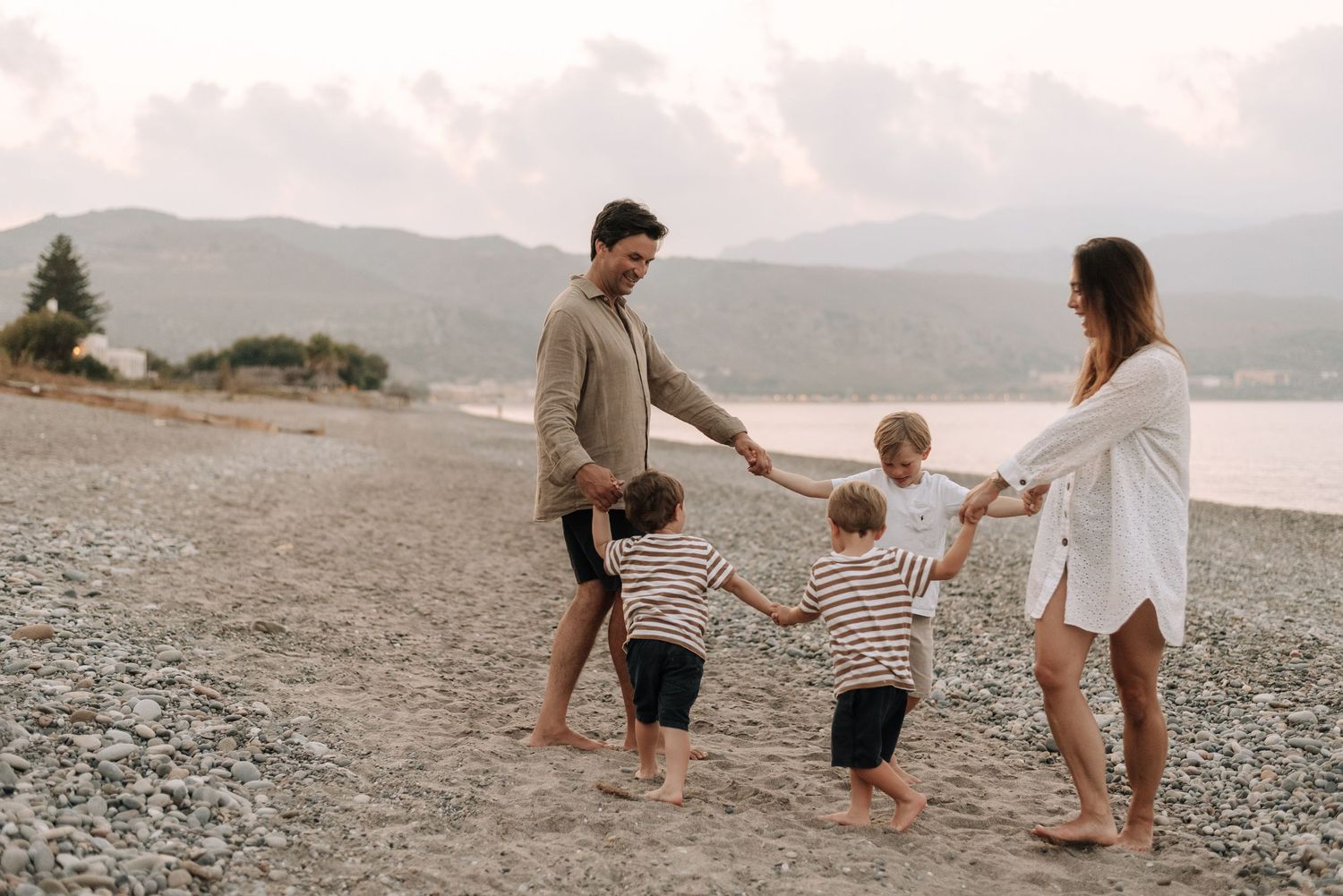 Happy family of four holding hands and walking together on a pebble beach with mountains in background.