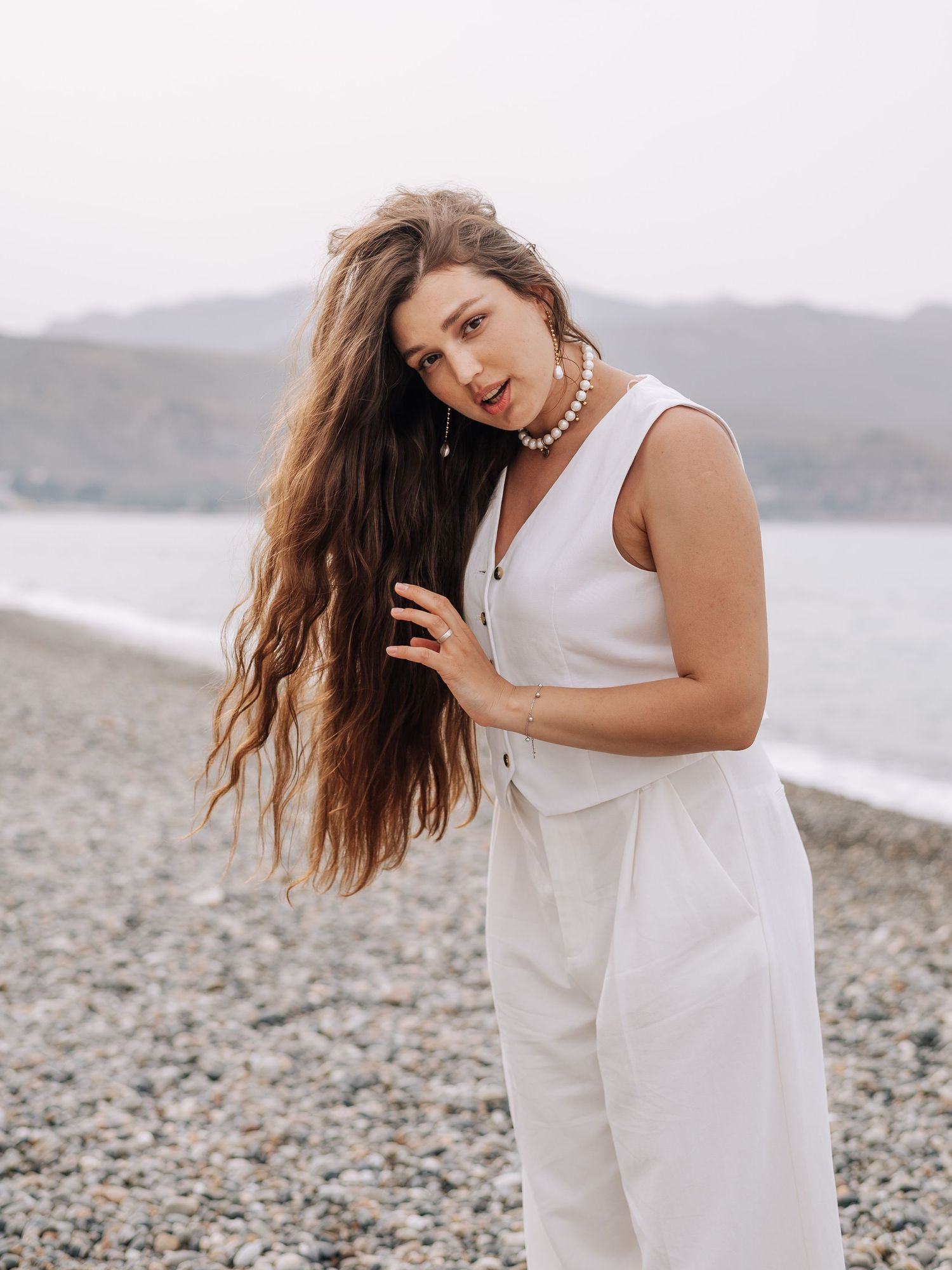 Woman with long wavy hair in white jumpsuit and pearl necklace posing on pebble beach.