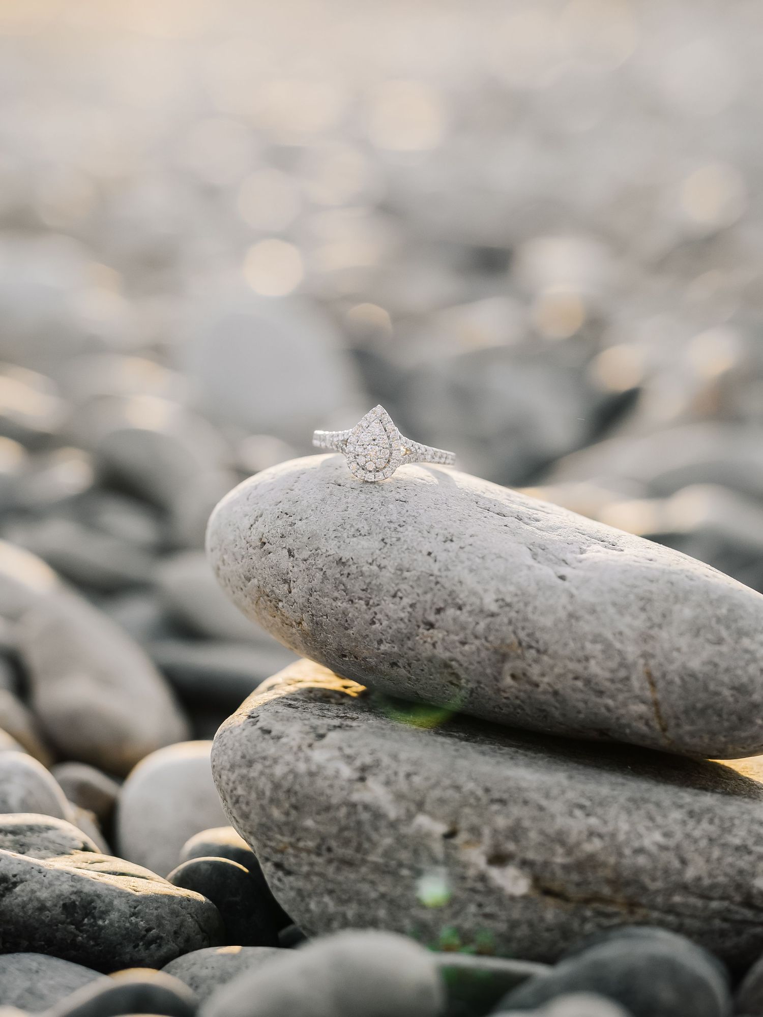 Diamond pear-shaped engagement ring balanced on stacked pebbles at golden hour on rocky beach.