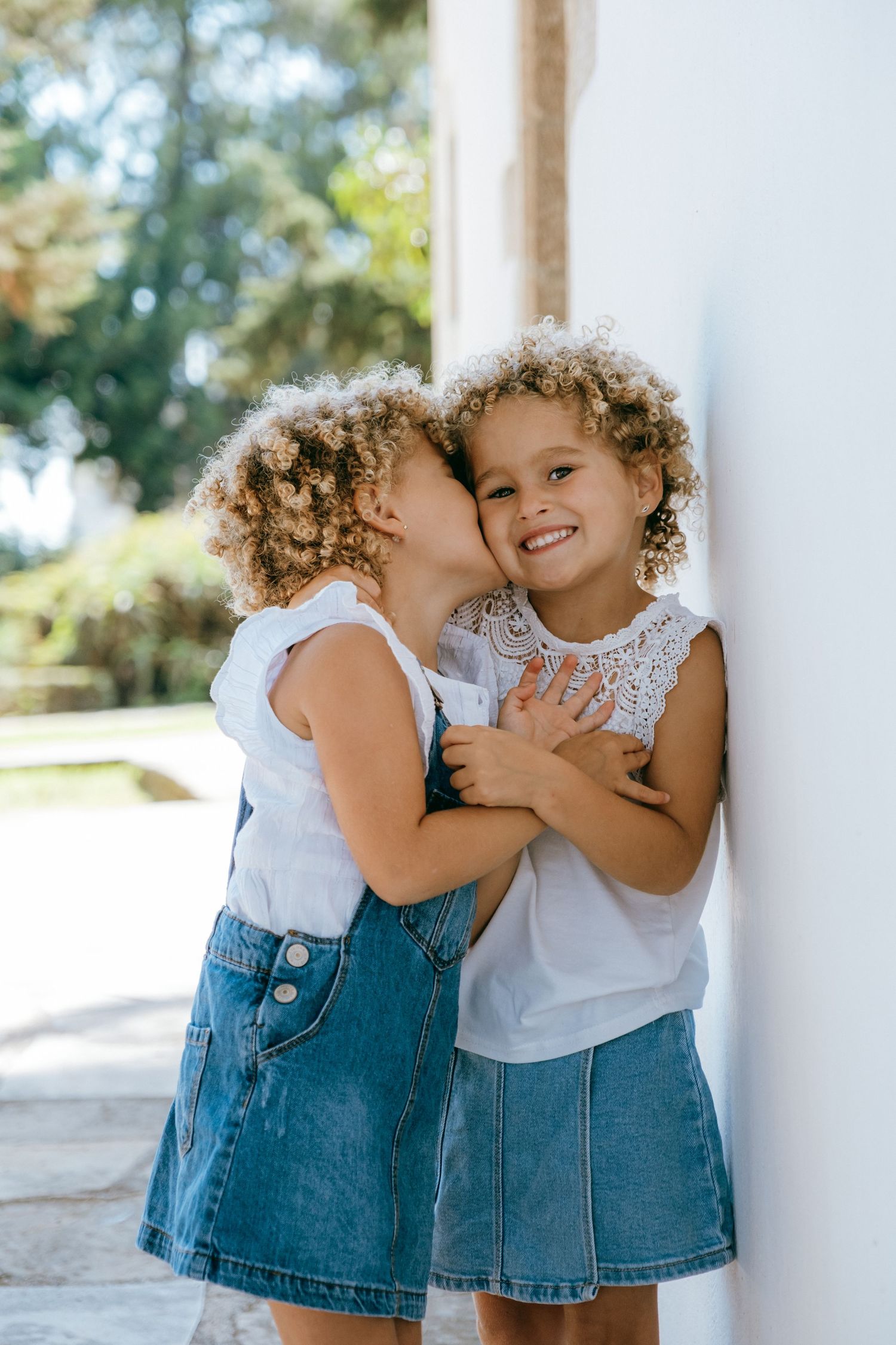 Twin girls with curly hair sharing a kiss on the cheek against a white wall.