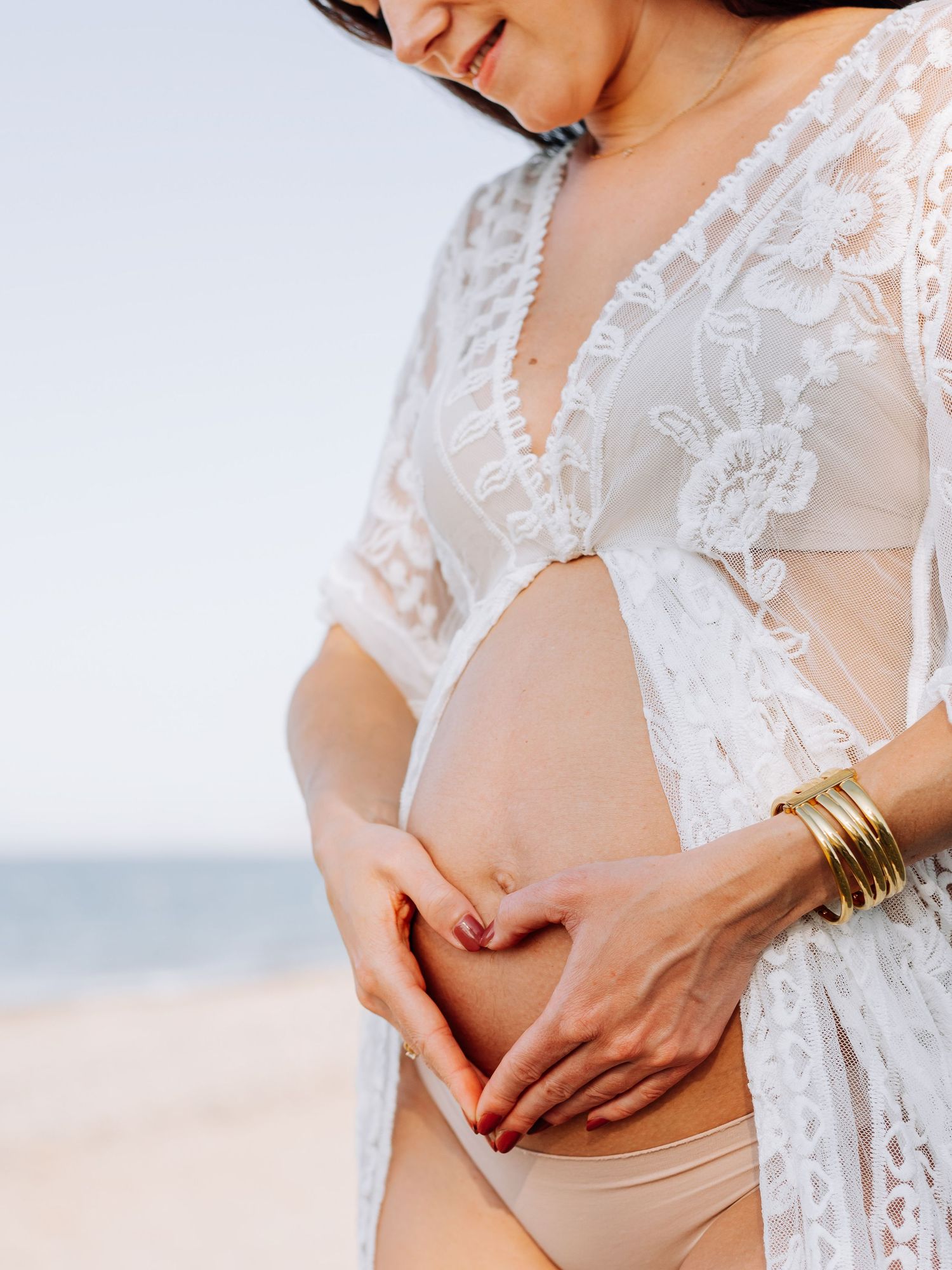 Pregnant woman in white lace robe forming heart shape with hands over bare baby bump at beach.