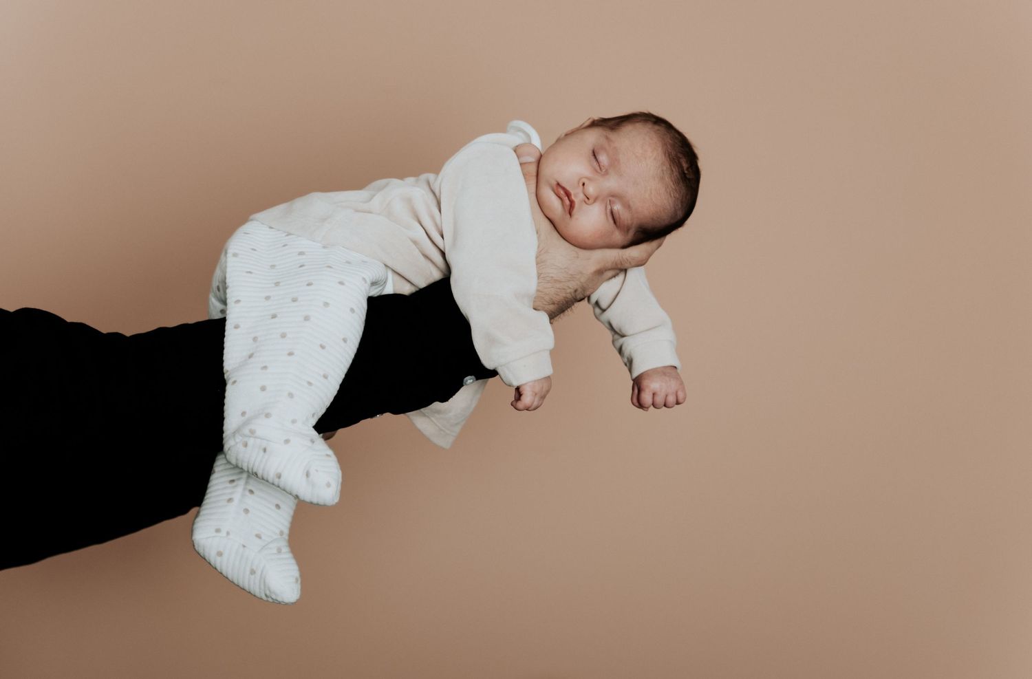 A peaceful sleeping newborn baby in a white onesie resting on a parent's arm against a beige background.