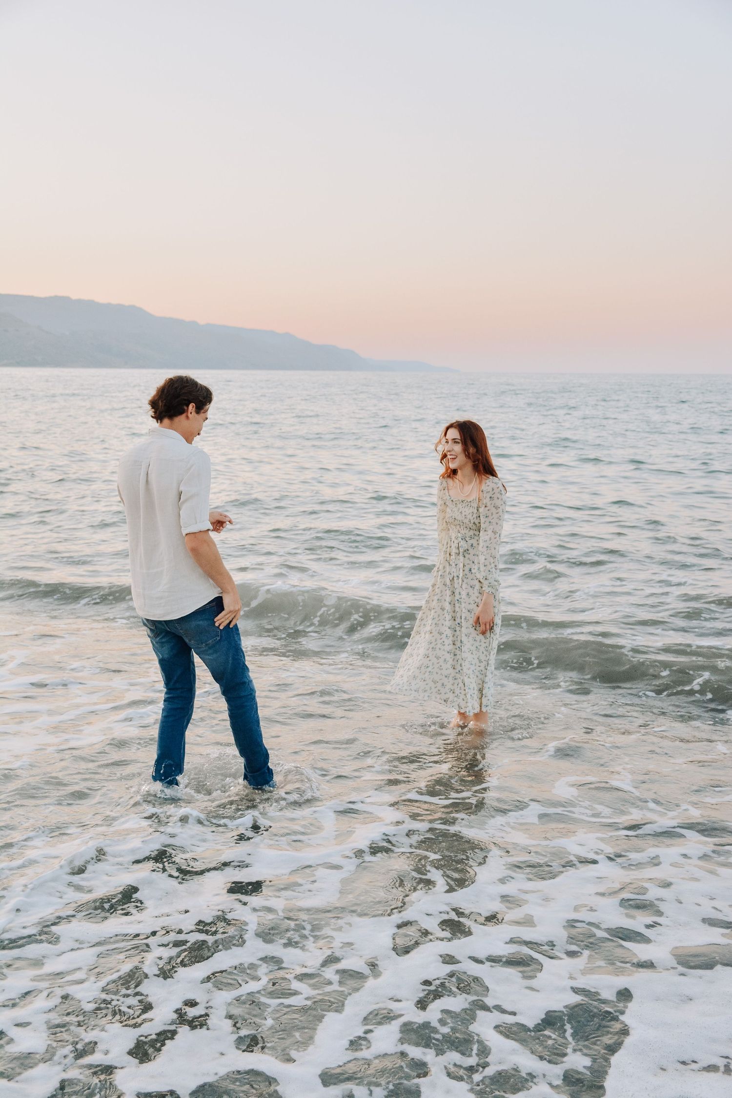 Couple standing in ocean waves at sunset, woman in floral dress and man in jeans sharing a romantic moment by the sea.