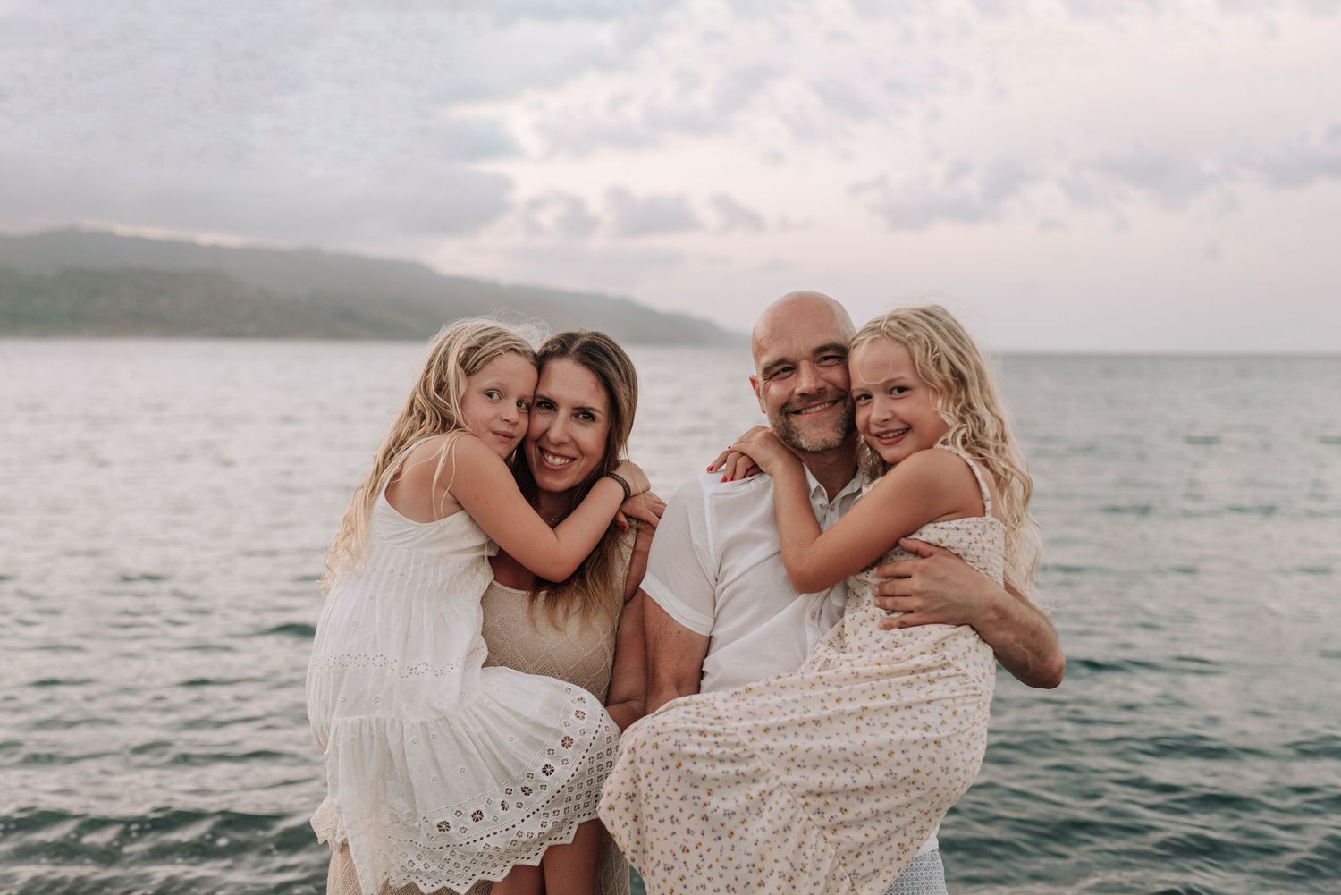 Family of four smiling together on a beach, parents holding two blonde girls in white dresses with ocean backdrop.