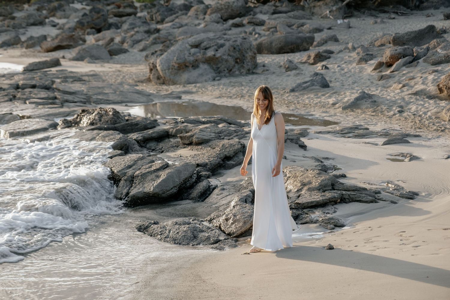 Bride in white wedding dress standing elegantly on sandy beach surrounded by coastal rocks.