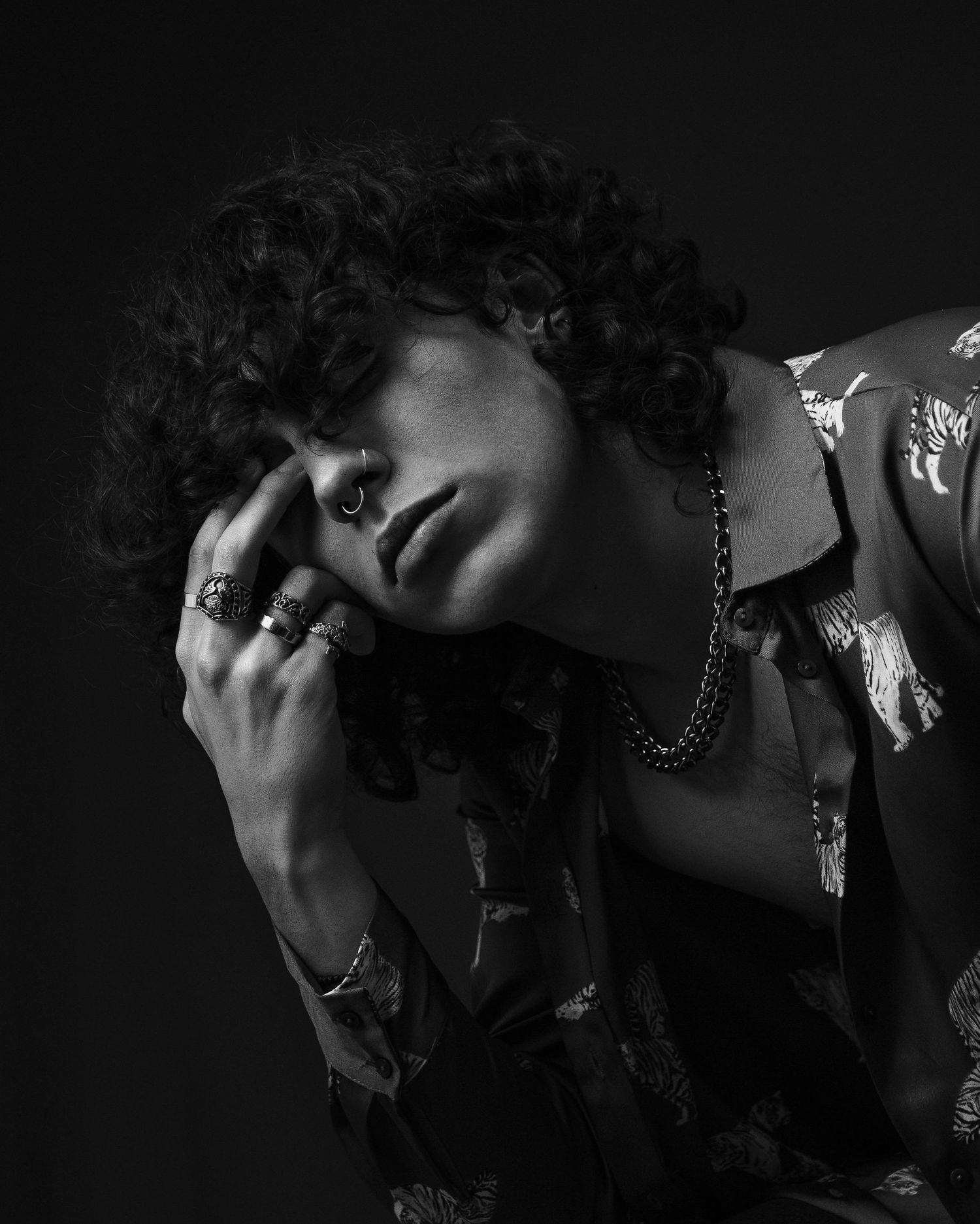 Black and white portrait of curly-haired person with rings and chain necklace in dramatic pose.