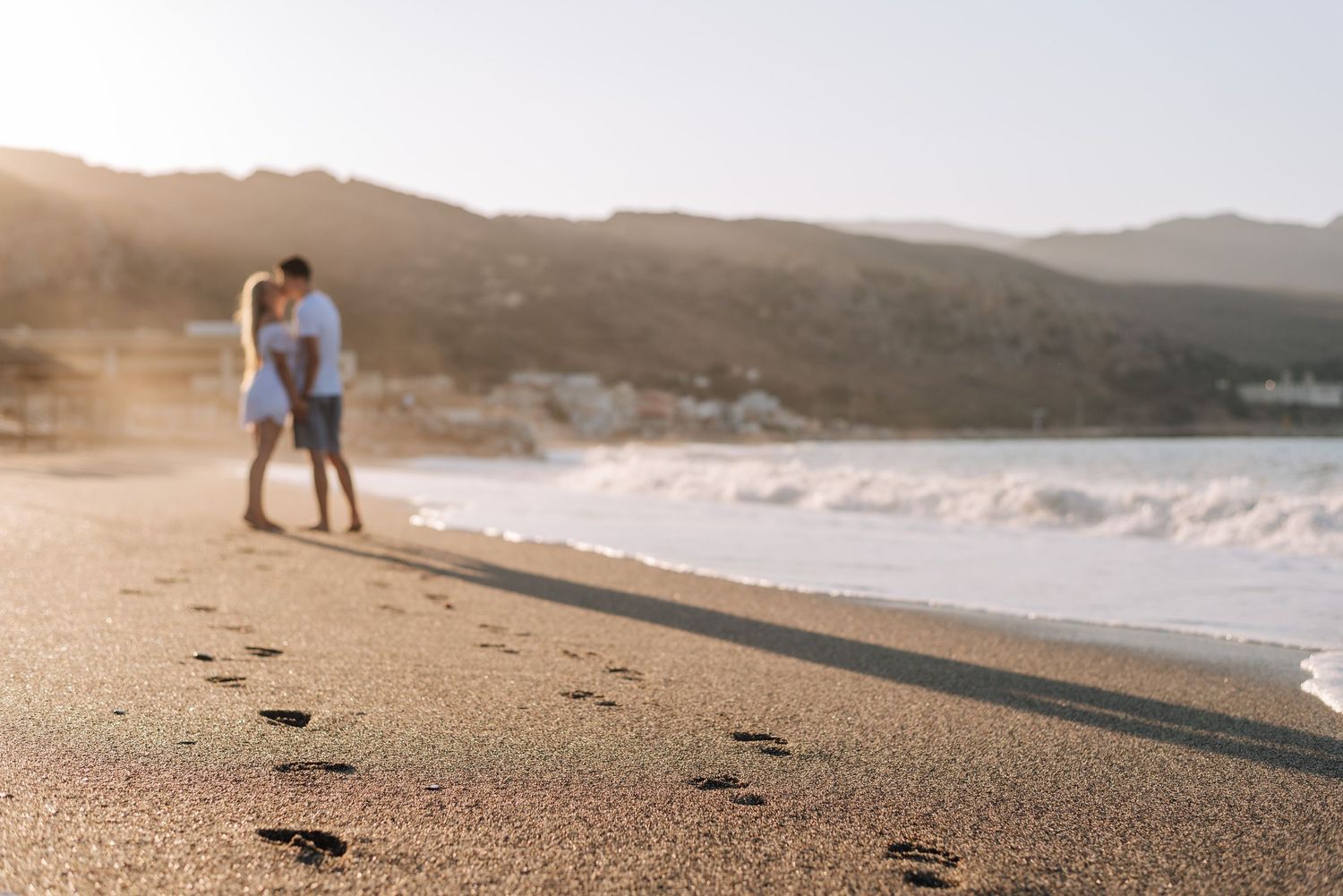 Couple walking hand in hand on a sandy beach at sunset, leaving footprints in the sand with ocean waves nearby.
