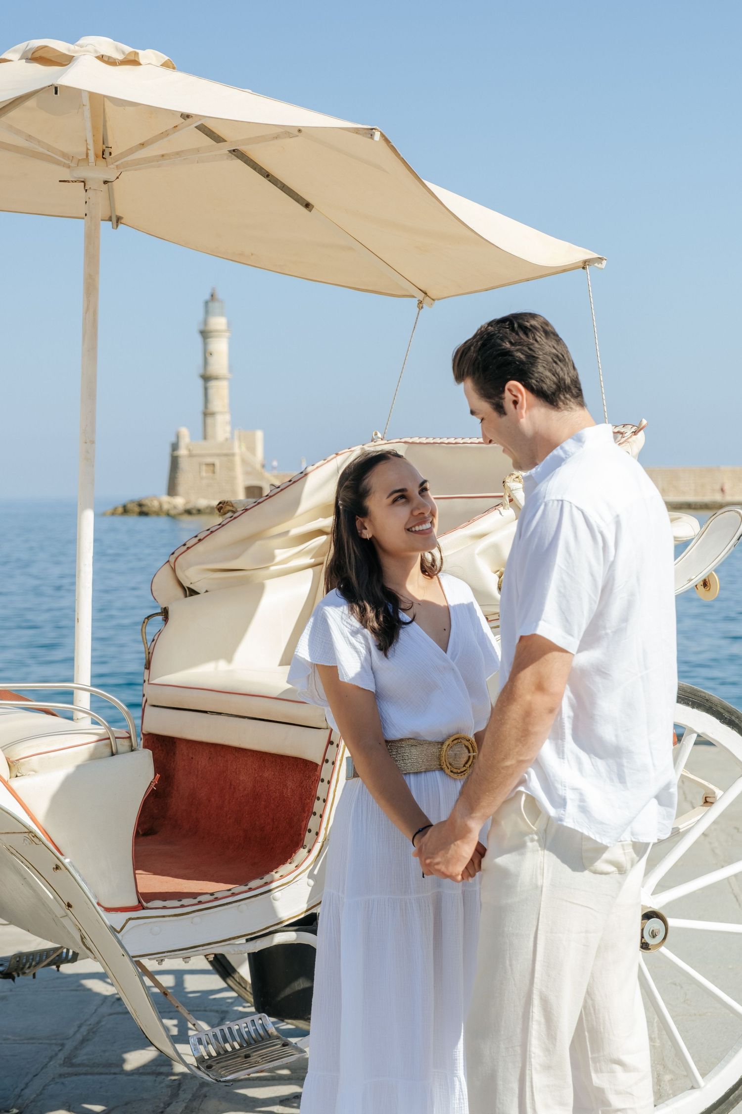 Romantic couple in white attire holds hands near vintage carriage with lighthouse and sea backdrop.
