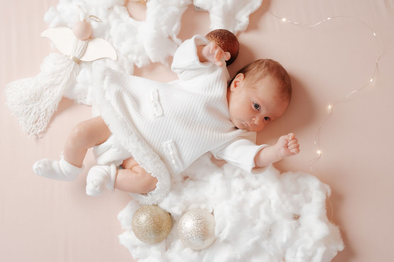 Newborn baby in white knit outfit lying on pink blanket surrounded by clouds and gold ornaments.