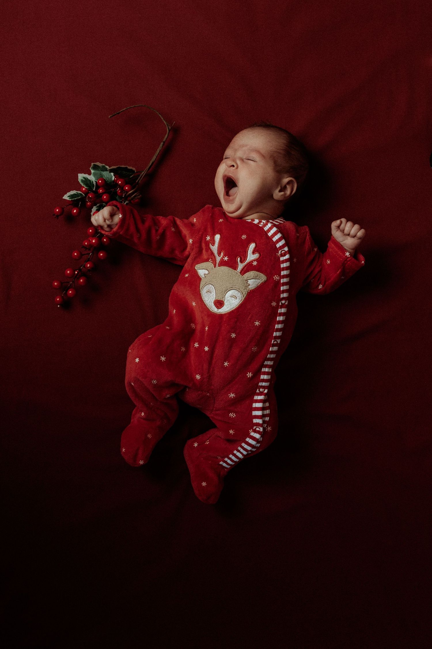 Newborn baby yawning in red reindeer Christmas onesie lying on red blanket with festive holly ornament.