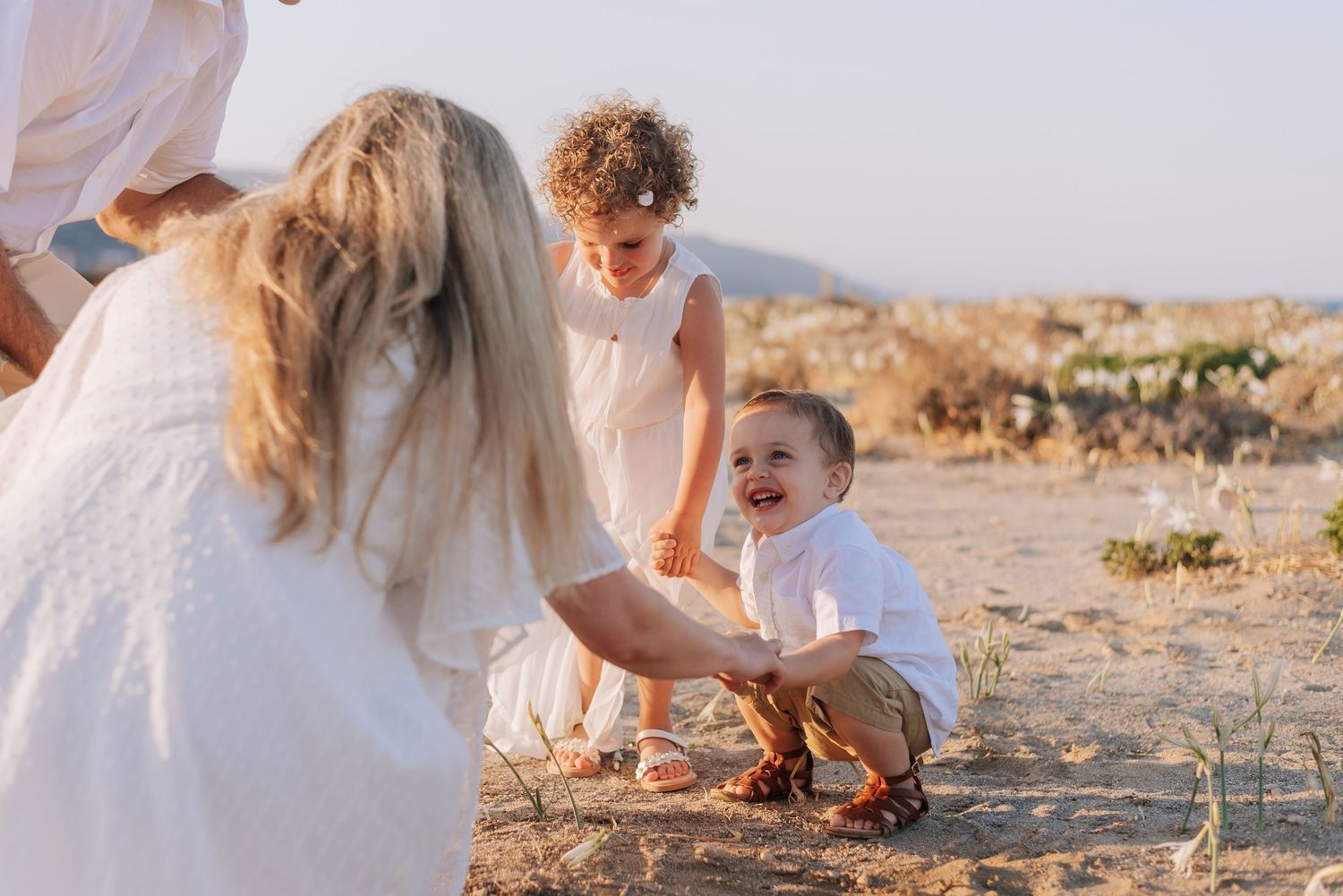 A happy toddler boy laughs while a woman reaches out to him as a child watches nearby outdoors.