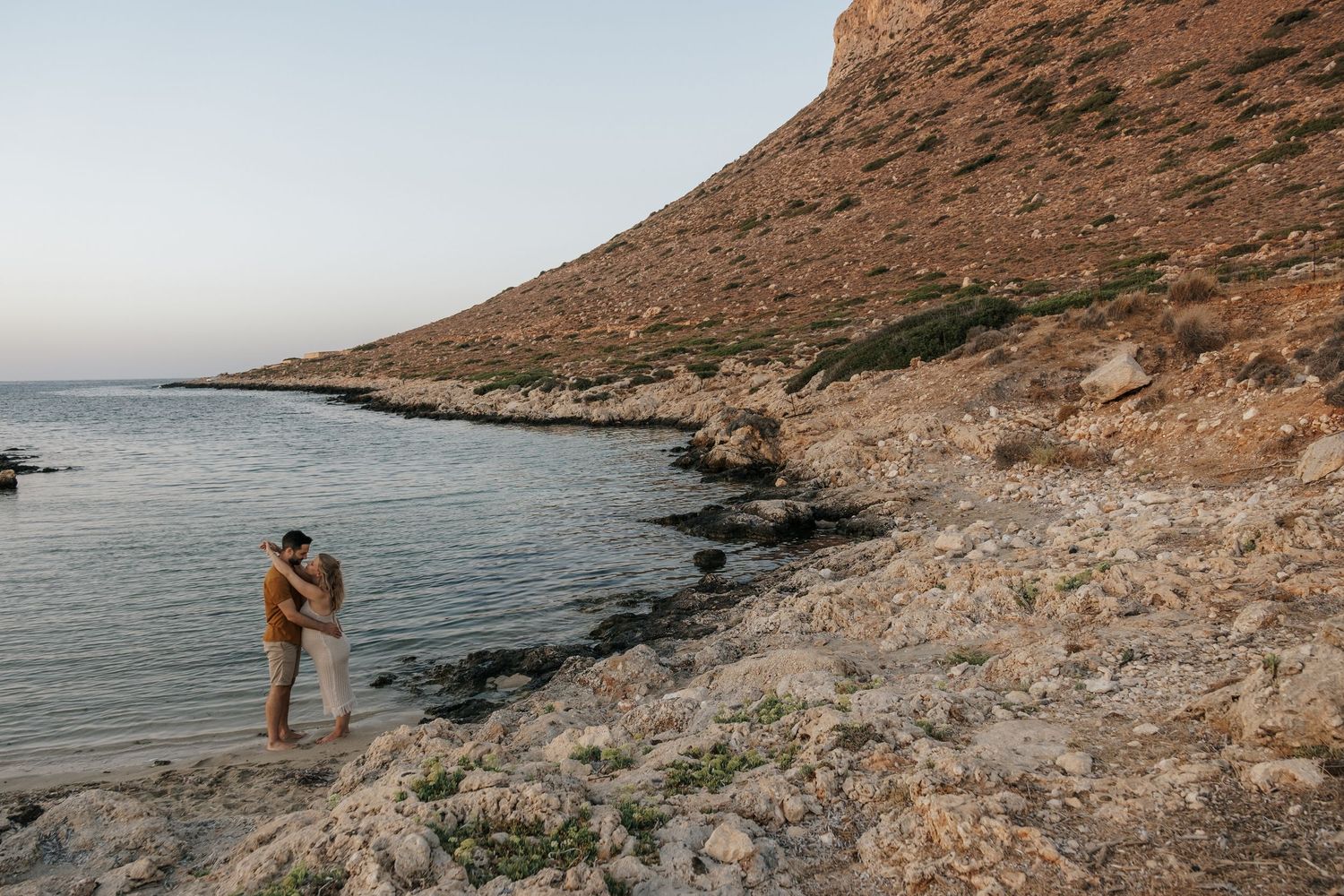 Couple sharing a romantic embrace on rocky coastal shore at sunset with calm blue waters.
