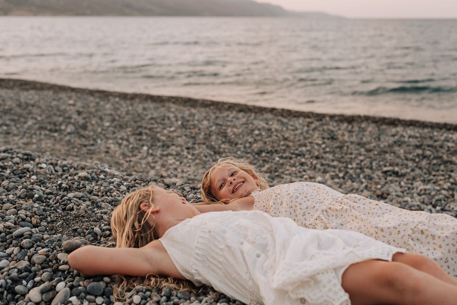Two girls in white dresses lying on a rocky beach laughing together.