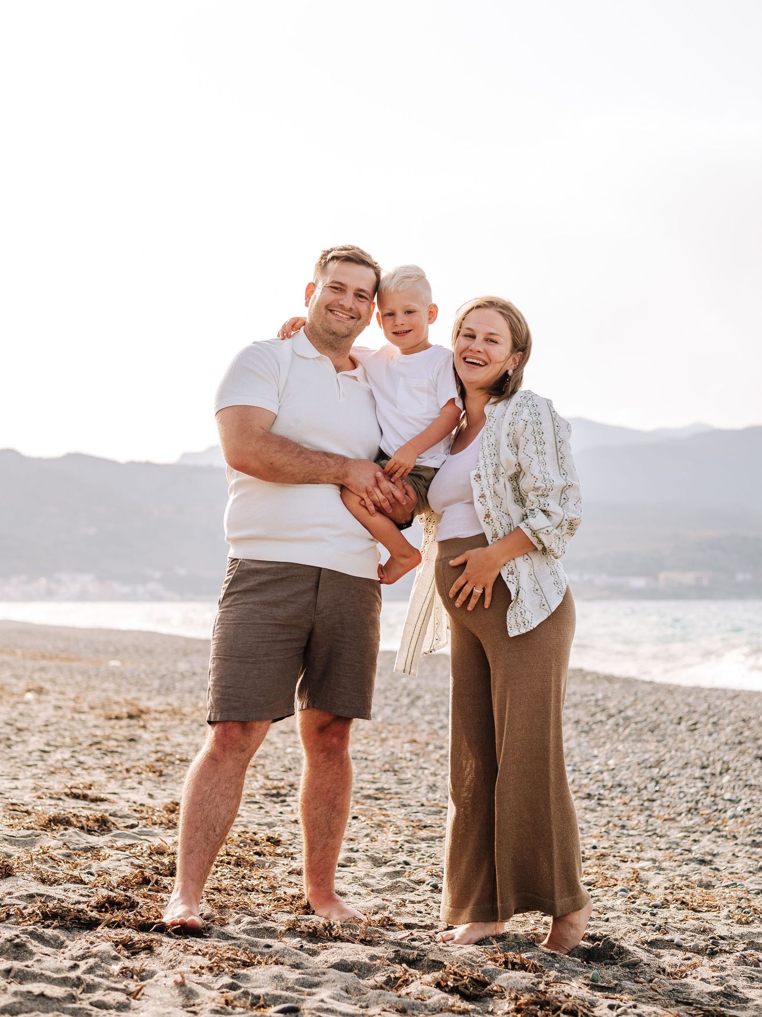 Happy family with toddler posing on sunny beach with pregnant mother and father smiling.