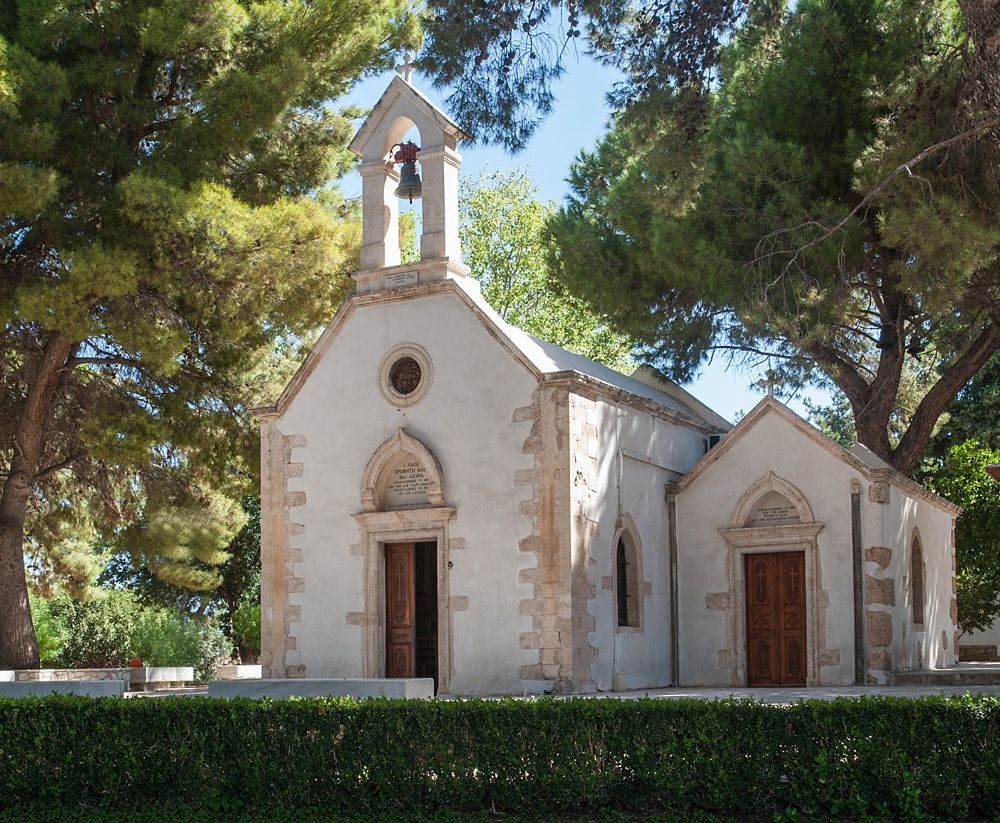 Small historic stone church with bell tower surrounded by tall pine trees under a clear blue sky.