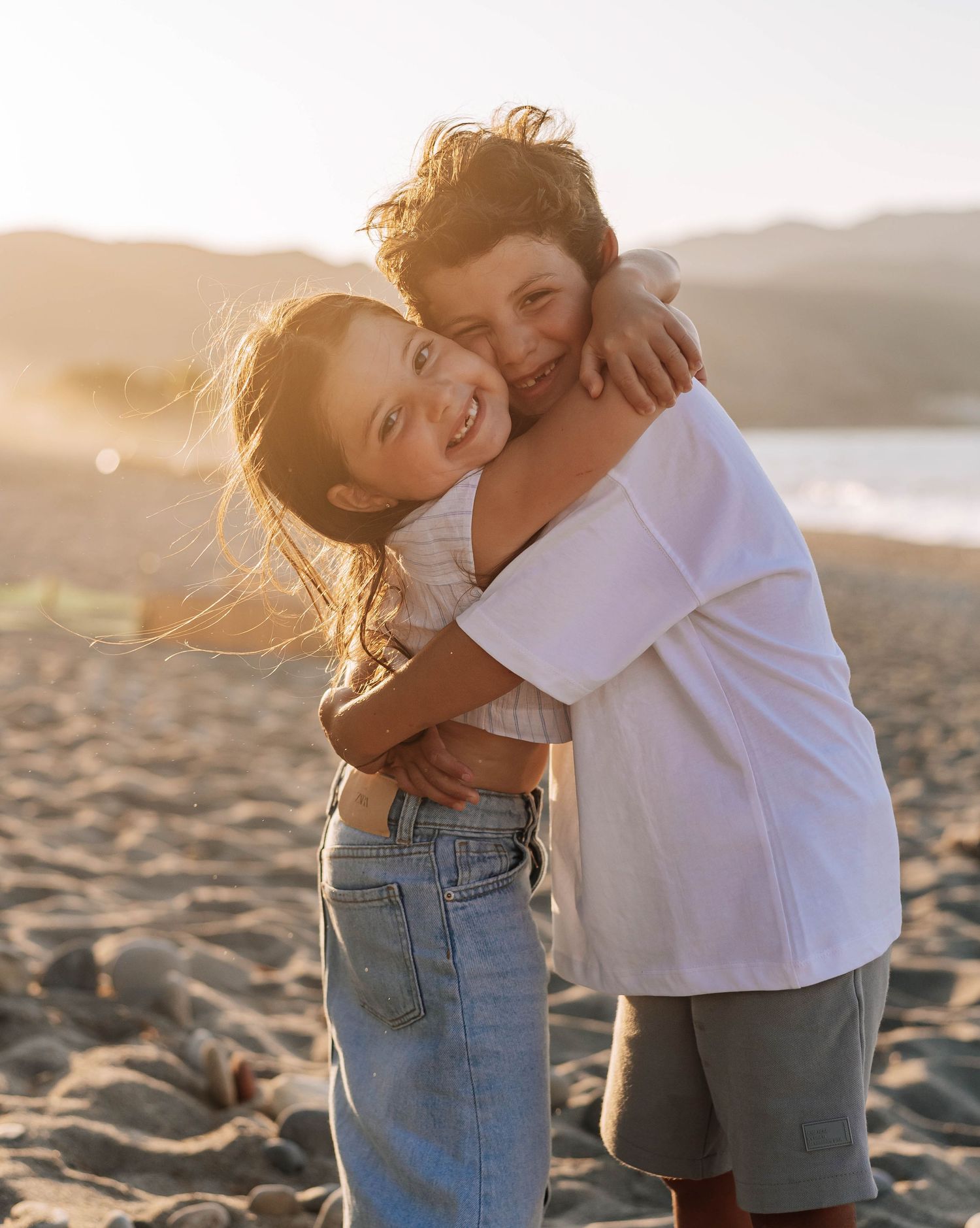 Two children hugging and smiling on a sunny beach at golden hour.
