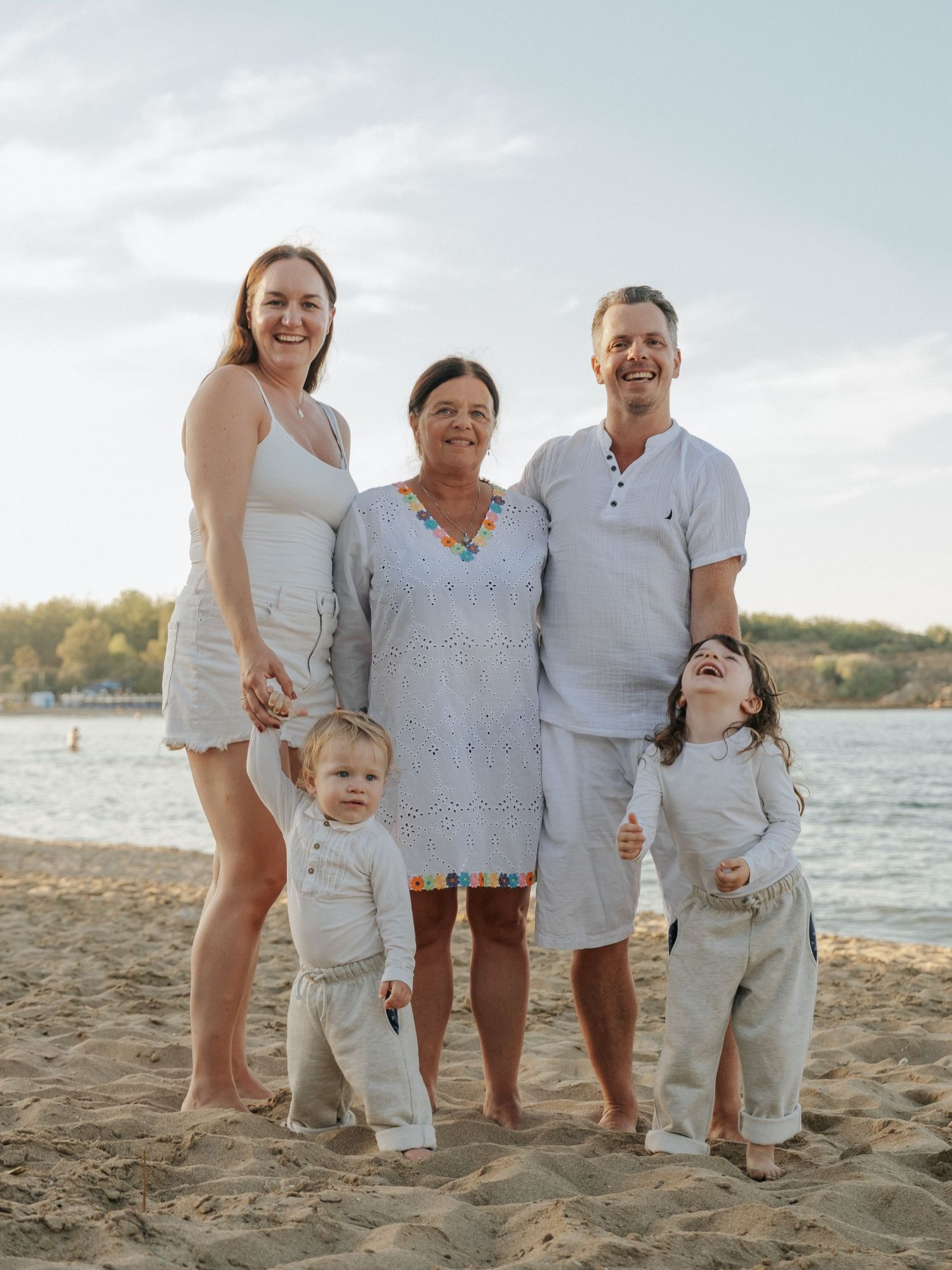 Family of five smiling together on sandy beach in coordinated white outfits.
