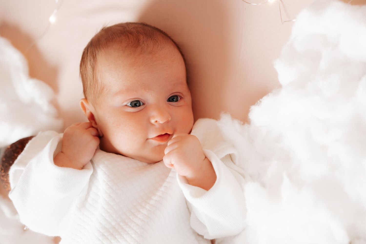 Adorable newborn baby in white outfit lying among fluffy white clouds with soft fairy lights.