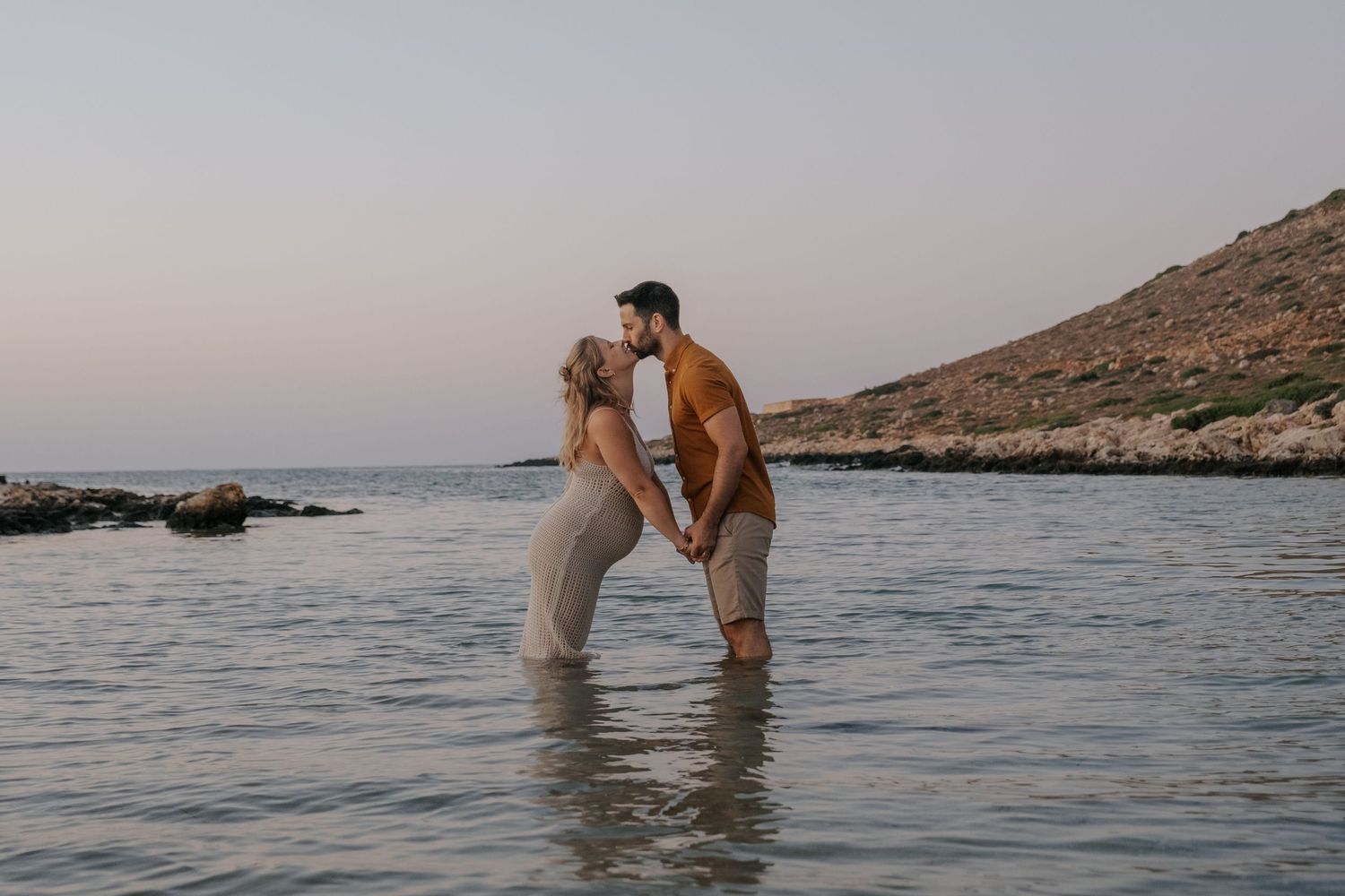 Romantic couple kissing while standing in shallow ocean water at dusk near rocky coastline.