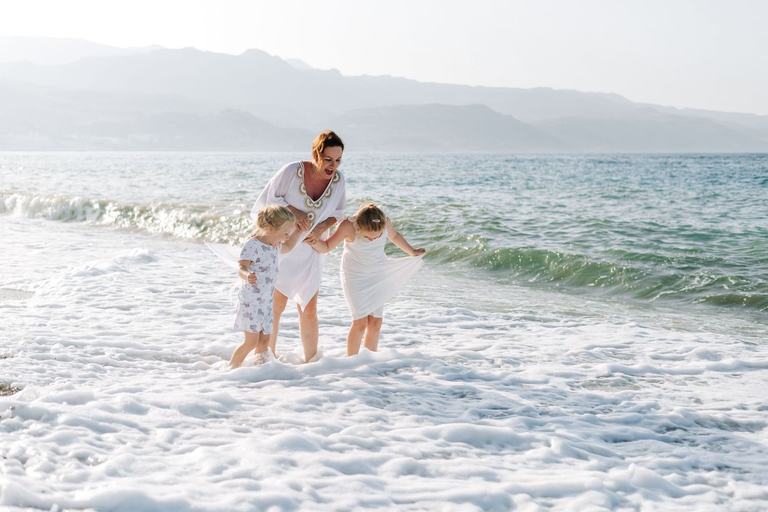 Mother playing with two young children in ocean waves on sunny beach.