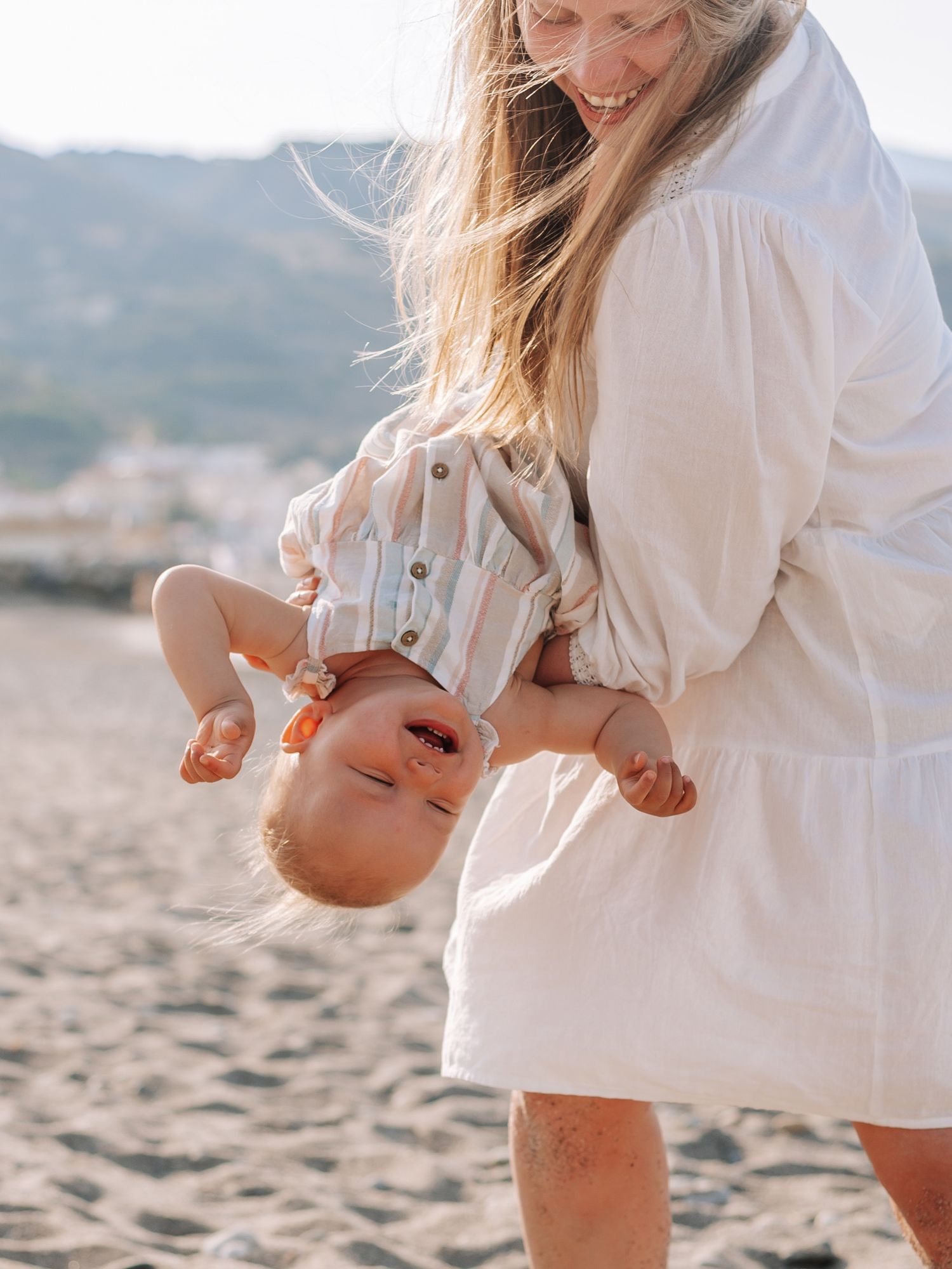 A joyful mother playfully holds her laughing baby upside down on a sunny beach.