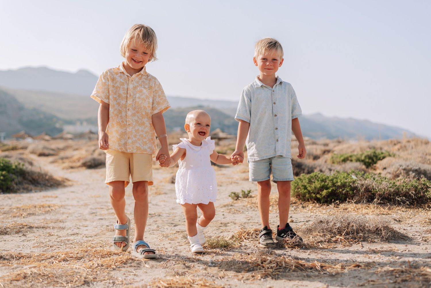 Three young siblings walk hand in hand along a scenic rocky path with mountains in the background.