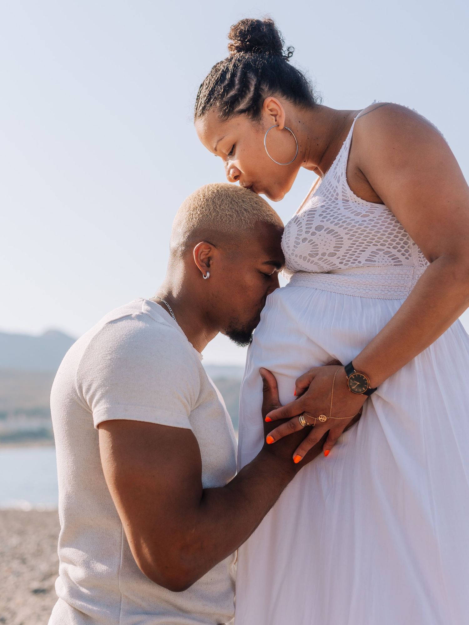 Man kneeling and kissing pregnant woman's belly on a sunny beach while she tenderly looks down at him.