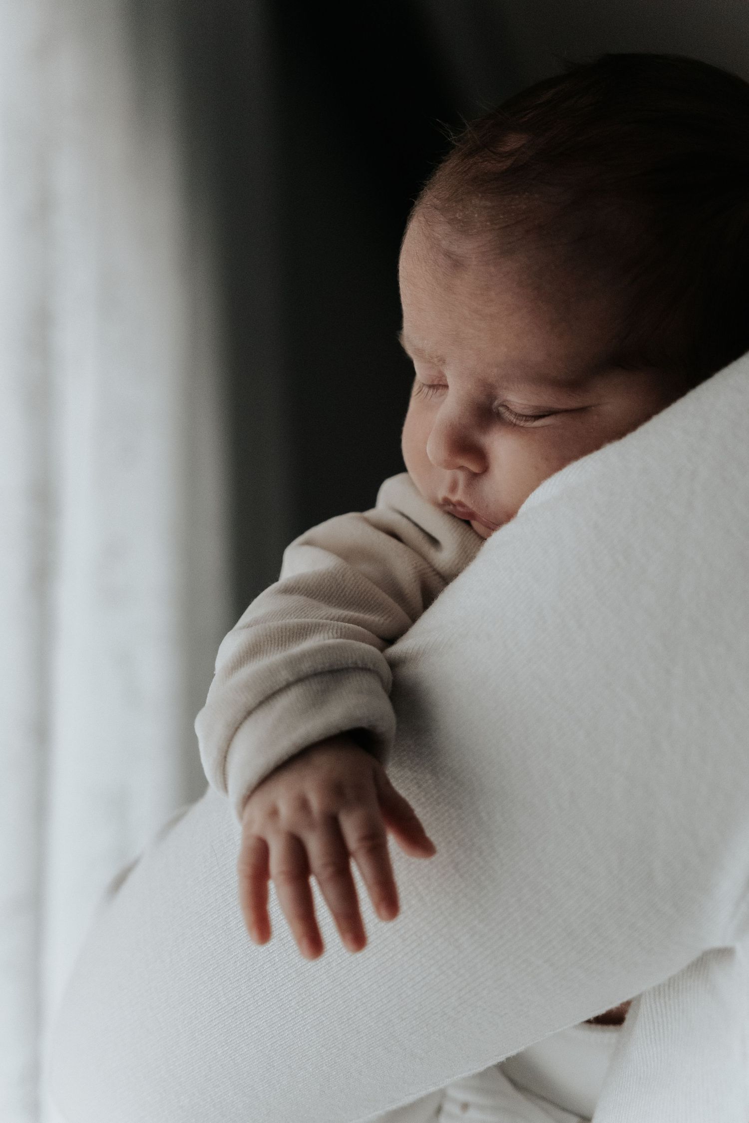 Peaceful sleeping newborn baby in beige outfit resting on a white cushion near a window.