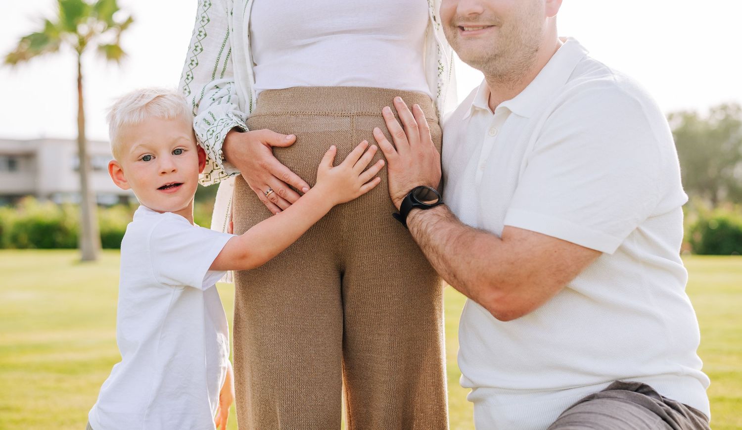 Young boy and father touching pregnant mothers belly outdoors in warm natural light.