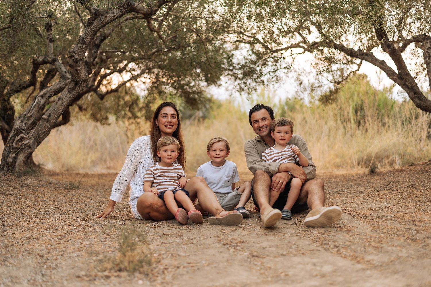 Happy family of five sitting together under olive trees in a sunny field.