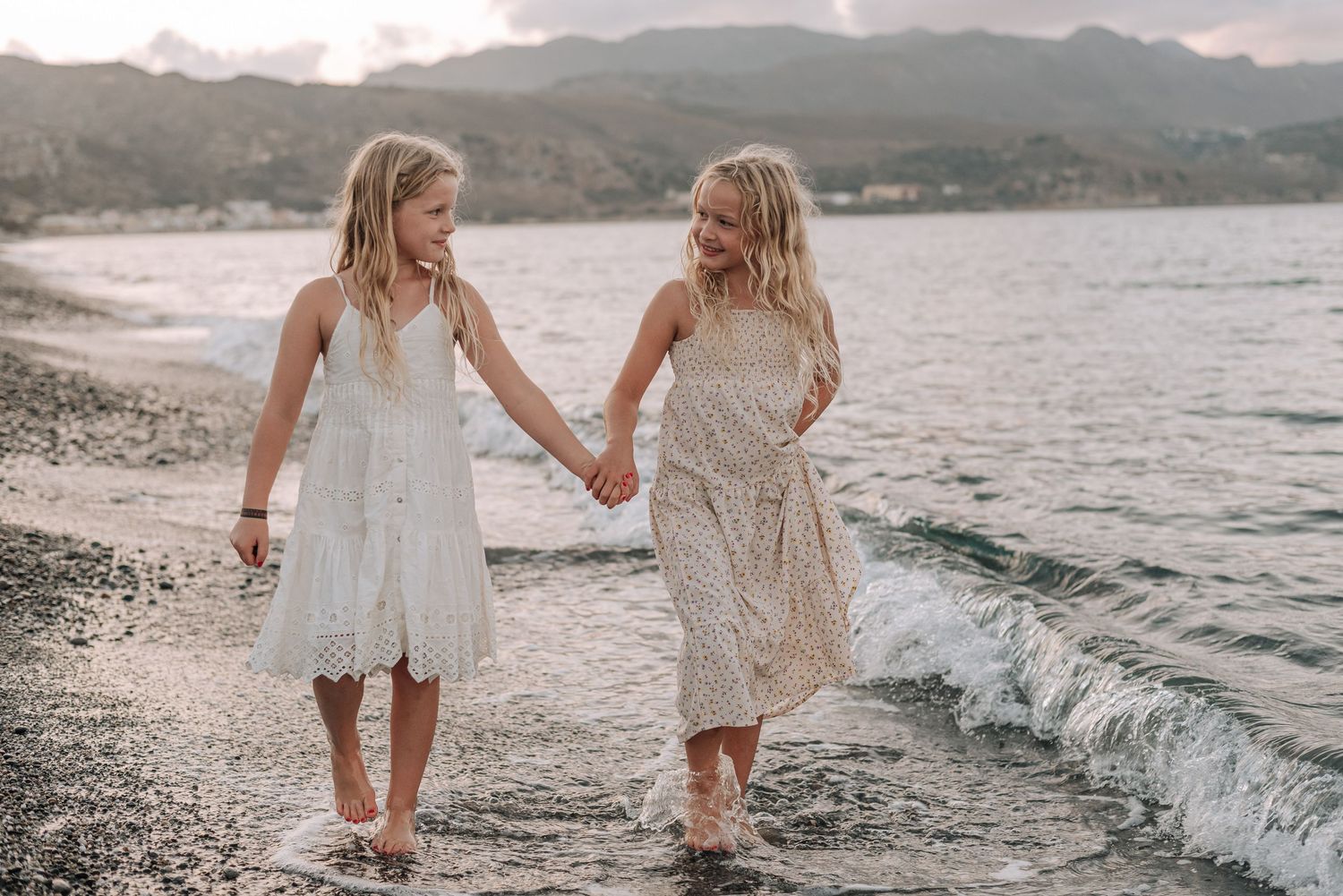 Two blonde girls in white dresses holding hands walking along ocean shoreline.