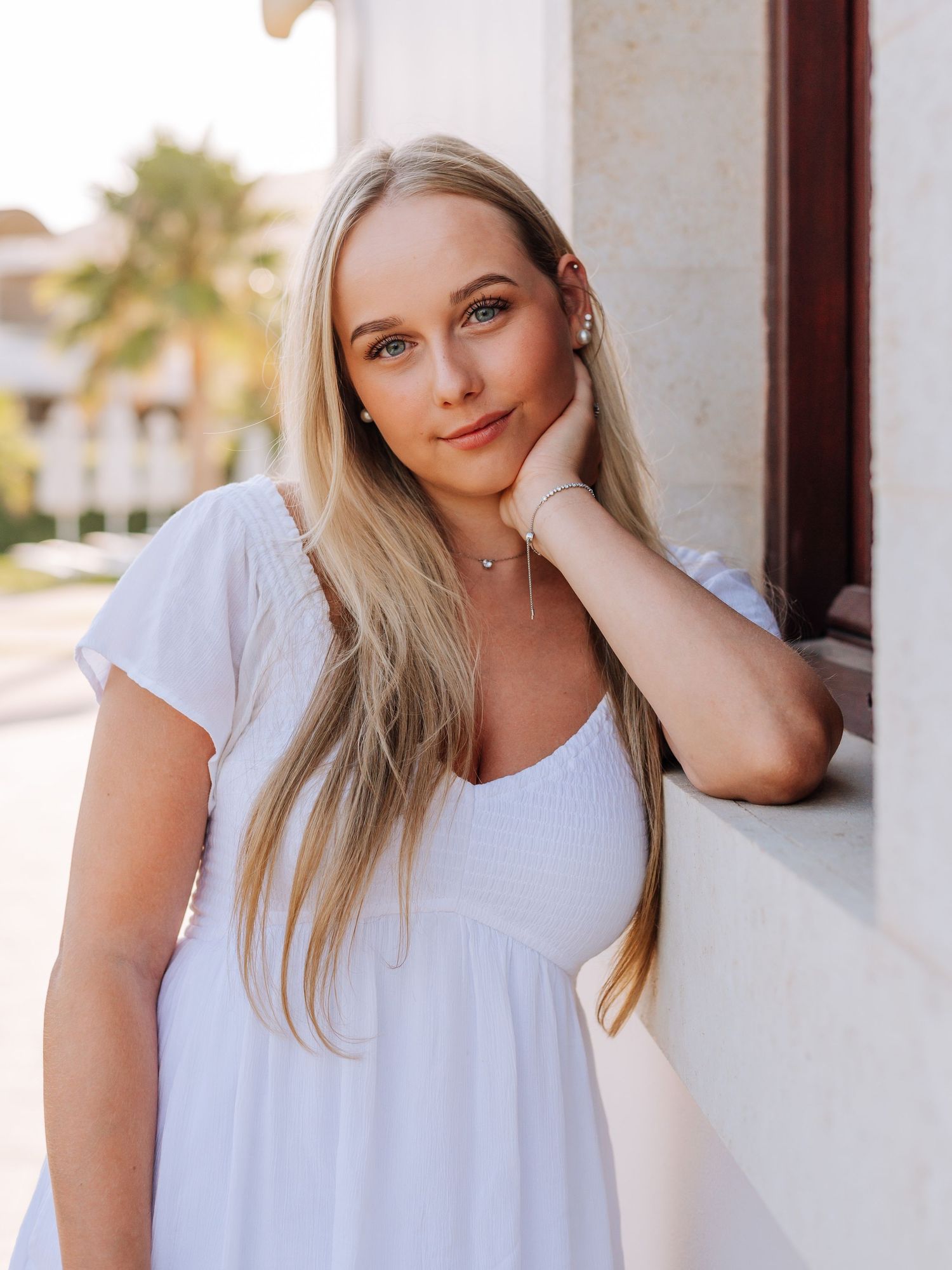 Blonde woman in white dress leaning against wall with palm trees in warm evening light.