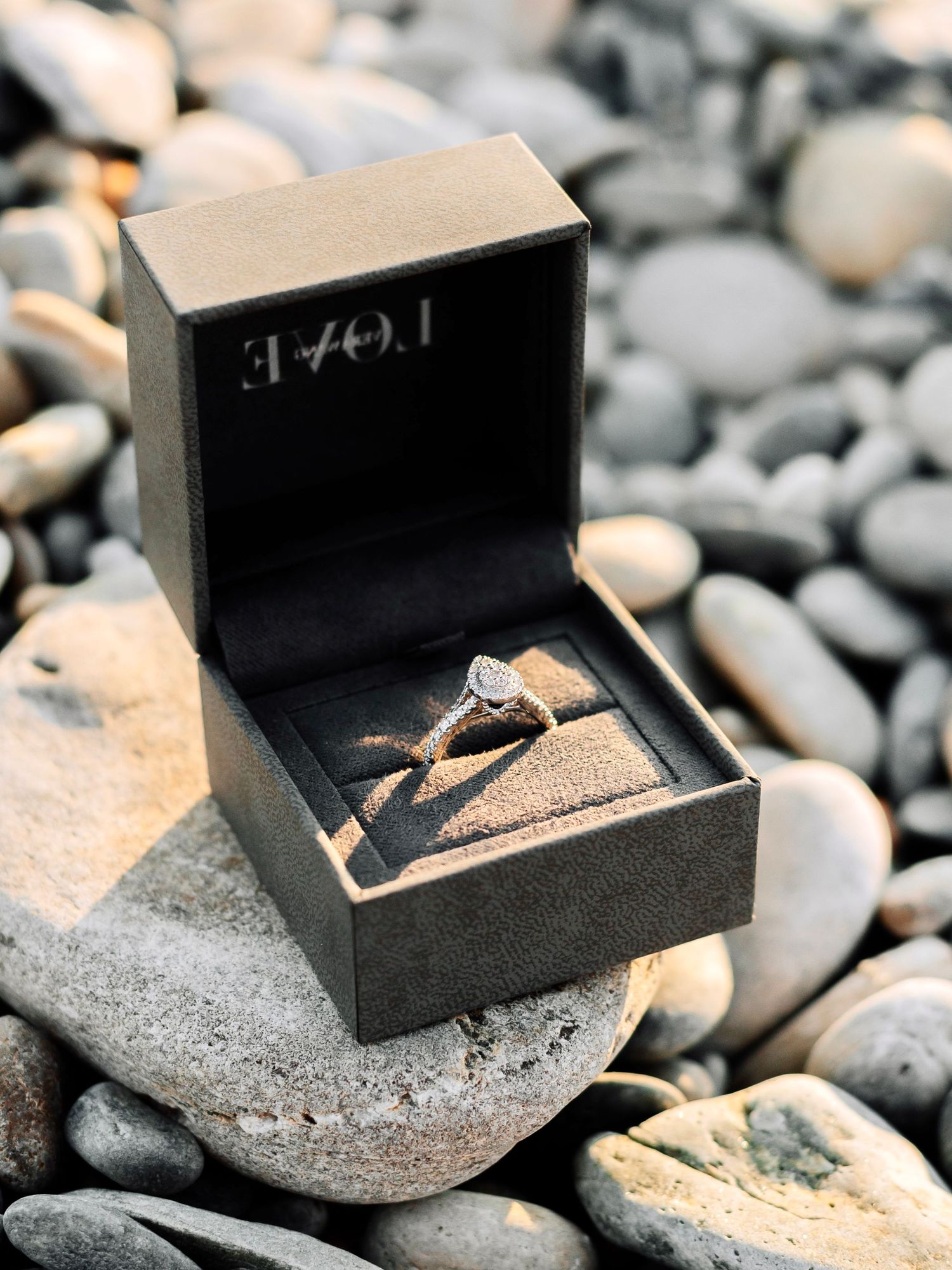 Diamond engagement ring displayed in an open black gift box on rocky beach.