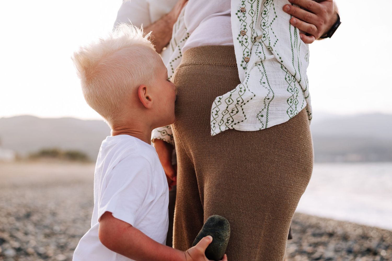 Young blonde toddler kissing pregnant mother's baby bump on a sunny beach.
