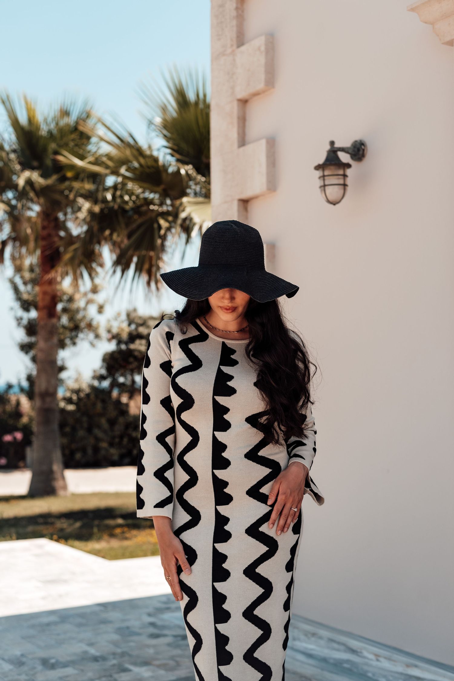 Woman in black and white zigzag patterned dress and wide-brim black hat posing by building with palm trees.