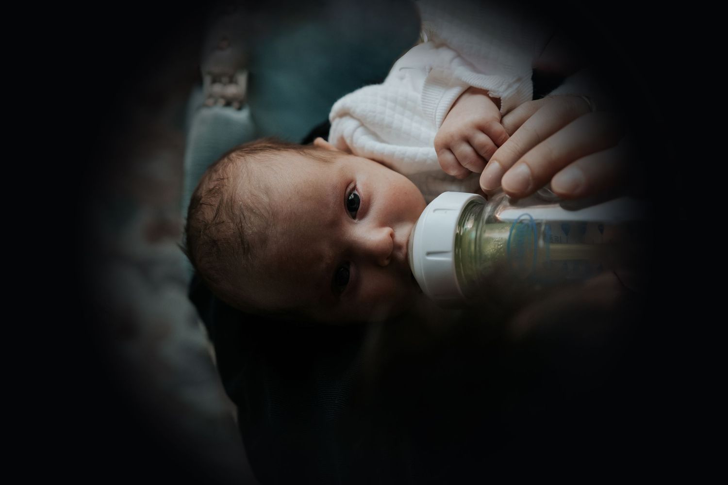 Newborn baby being bottle-fed in dim light, cradled gently in caregiver's arms.