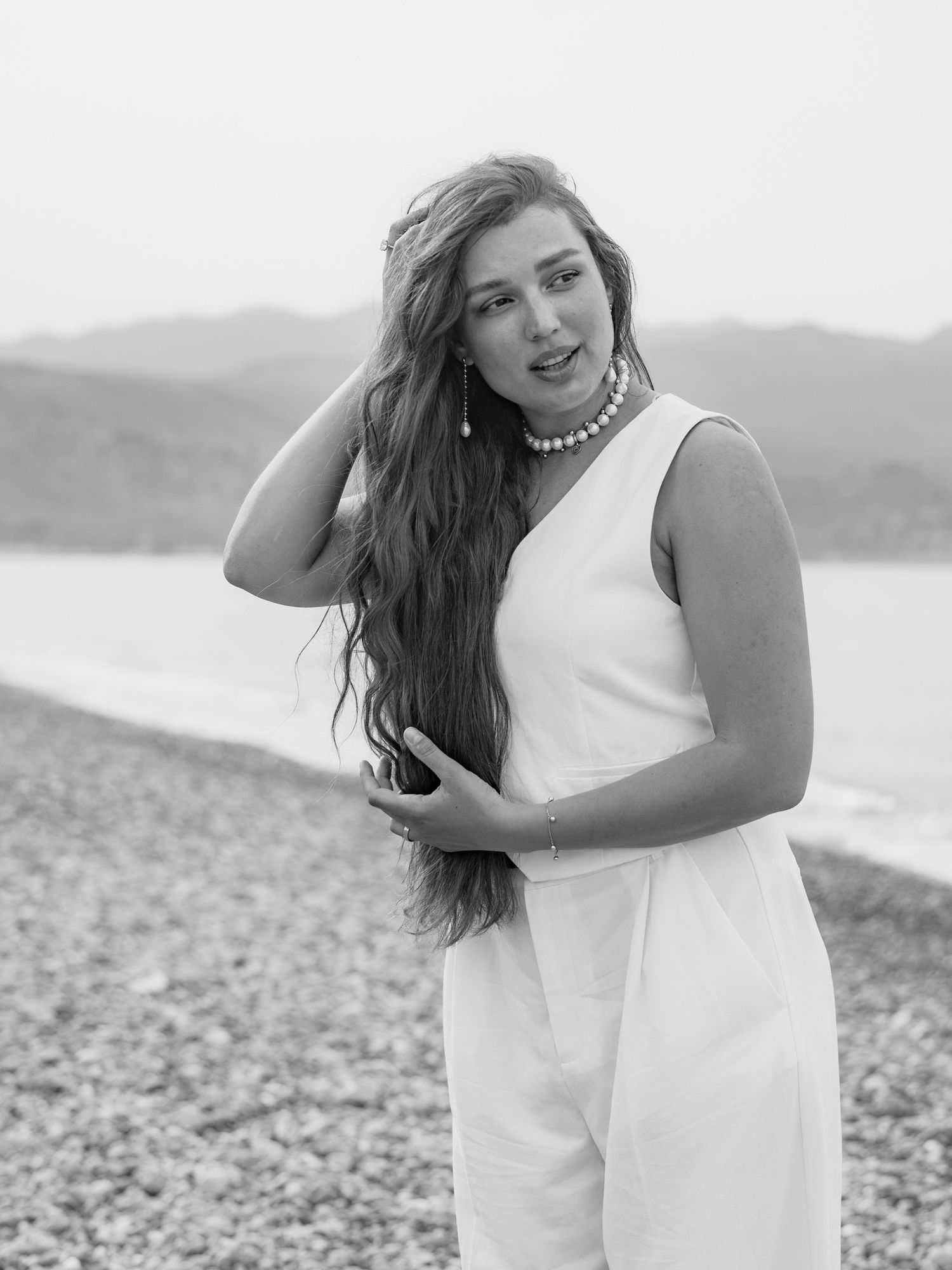 Young woman in white jumpsuit and pearl necklace posing on rocky beach in black and white photo.