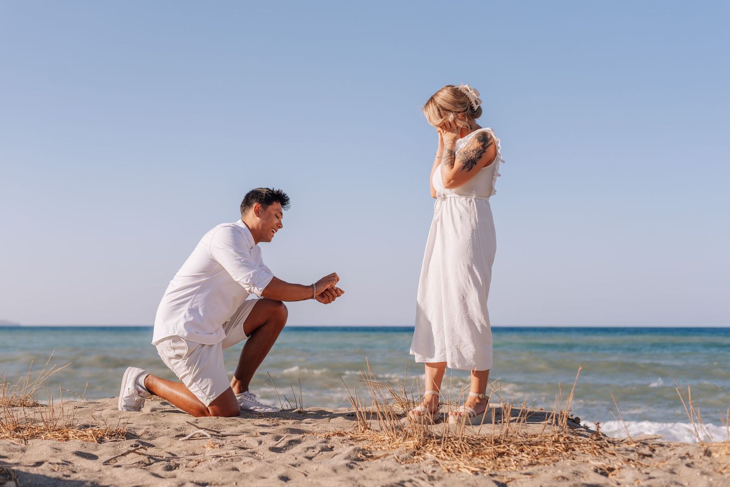 Man proposing on one knee to a woman in a white dress on a sunny sandy beach by the sea.
