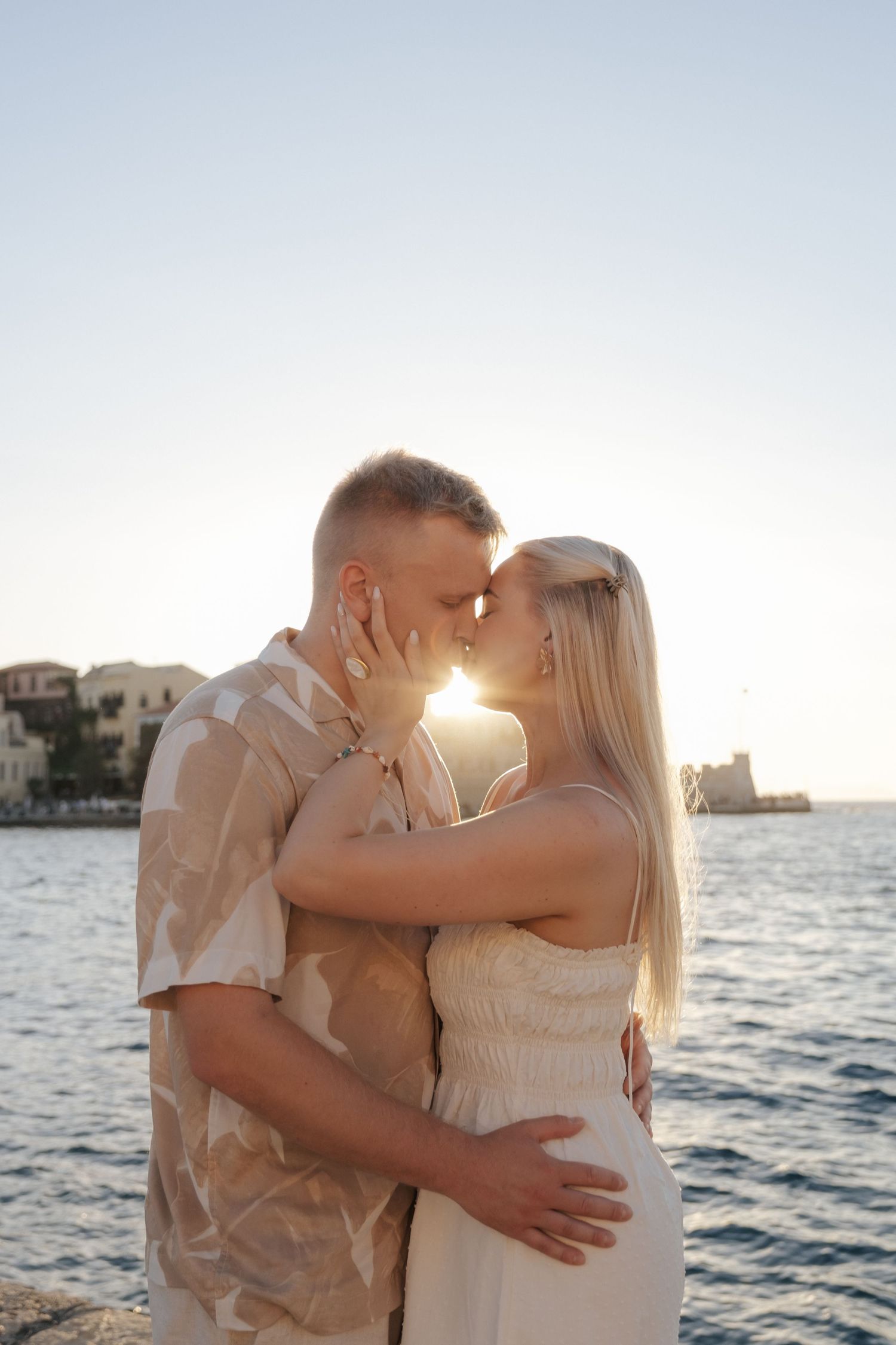 Couple shares romantic almost-kiss at sunset with sparkling sea and coastal town in background.