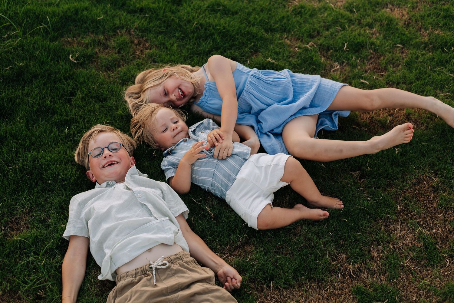 Three happy children laughing and relaxing together on green grass outdoors on a sunny day.