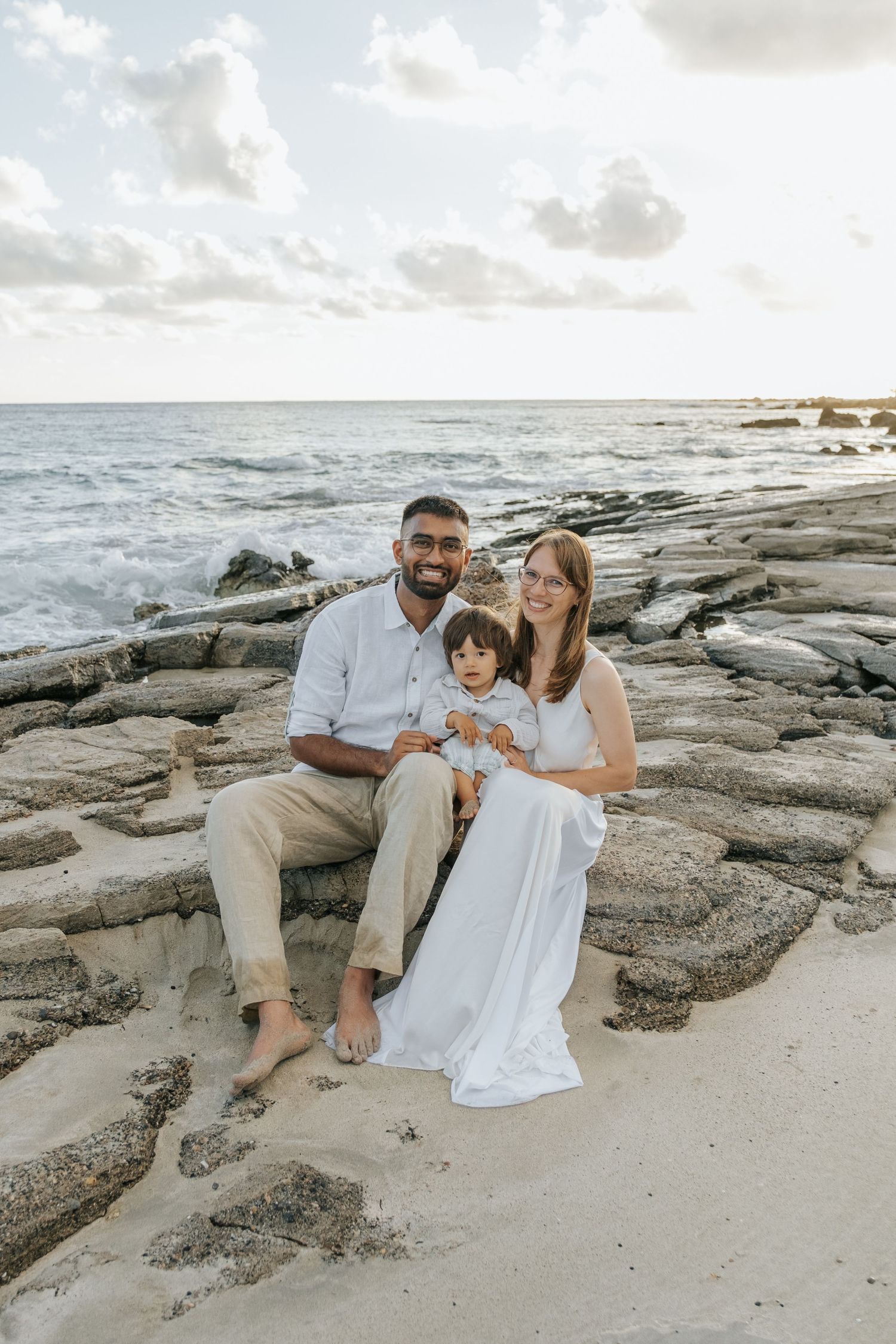Happy family with baby poses on rocky beach at sunset in coordinating white and tan outfits.