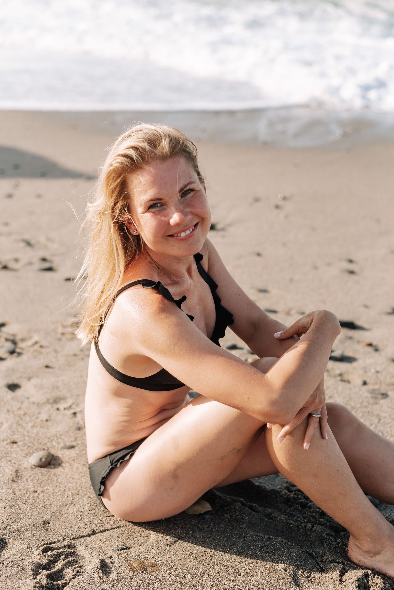 Smiling blonde woman in black bikini sitting relaxed on sandy beach with ocean waves behind.