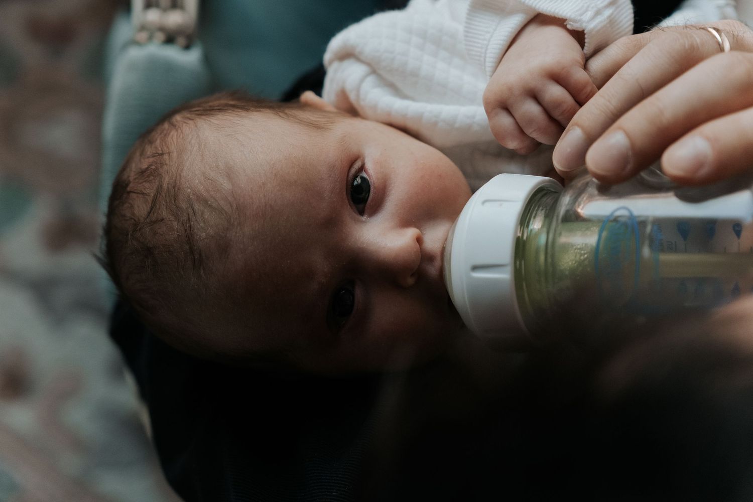 Newborn baby with dark hair drinking milk from a bottle, gazing upward with curious eyes.