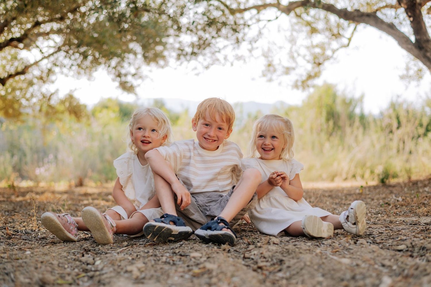 Three young blonde siblings sit together smiling under a tree in a sun-dappled outdoor setting.