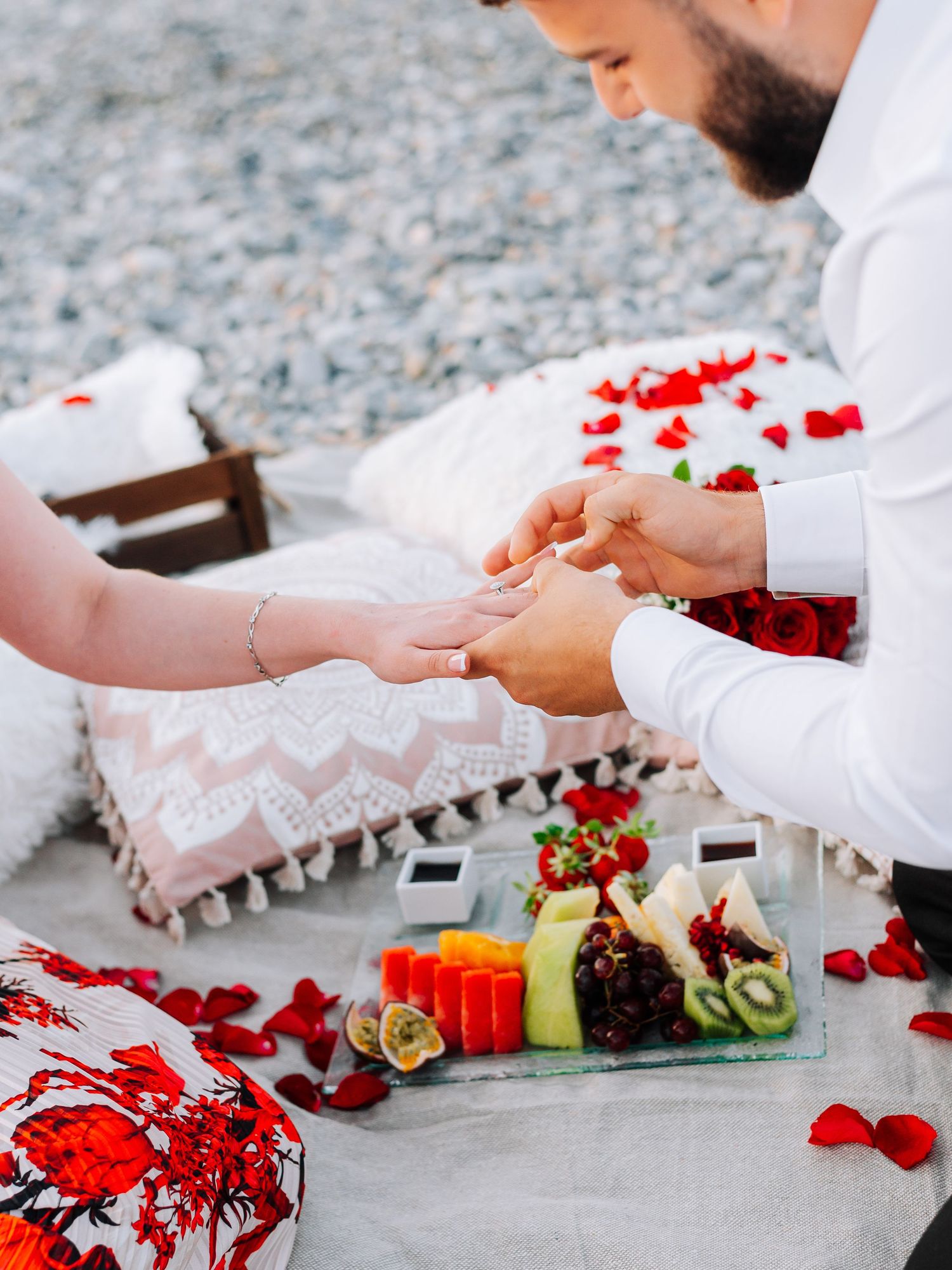 Man placing engagement ring on woman's finger during a romantic beach picnic with rose petals.